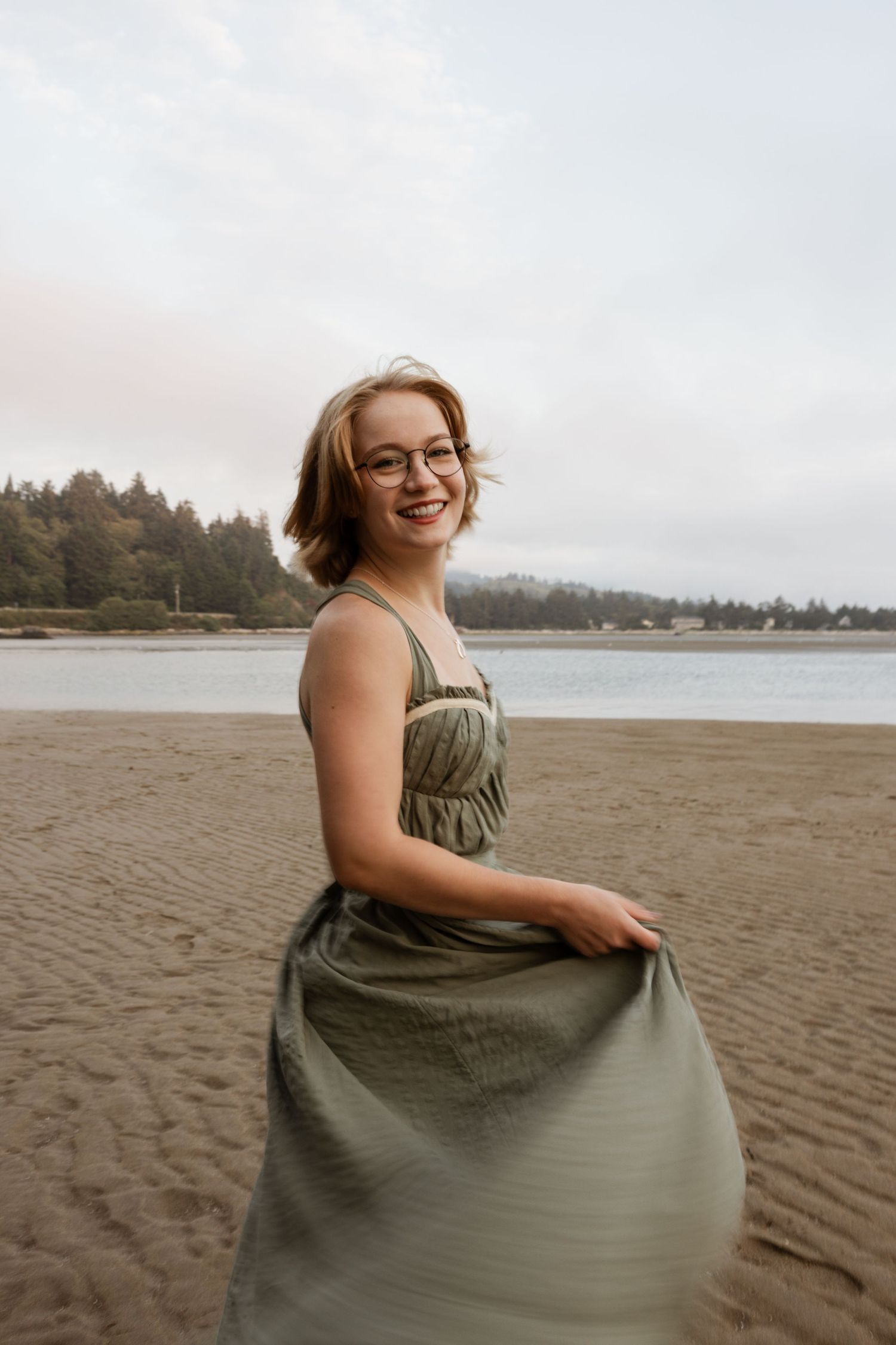 A person in a green dress smiles joyfully while sitting on a sandy beach with evergreen trees in the background.