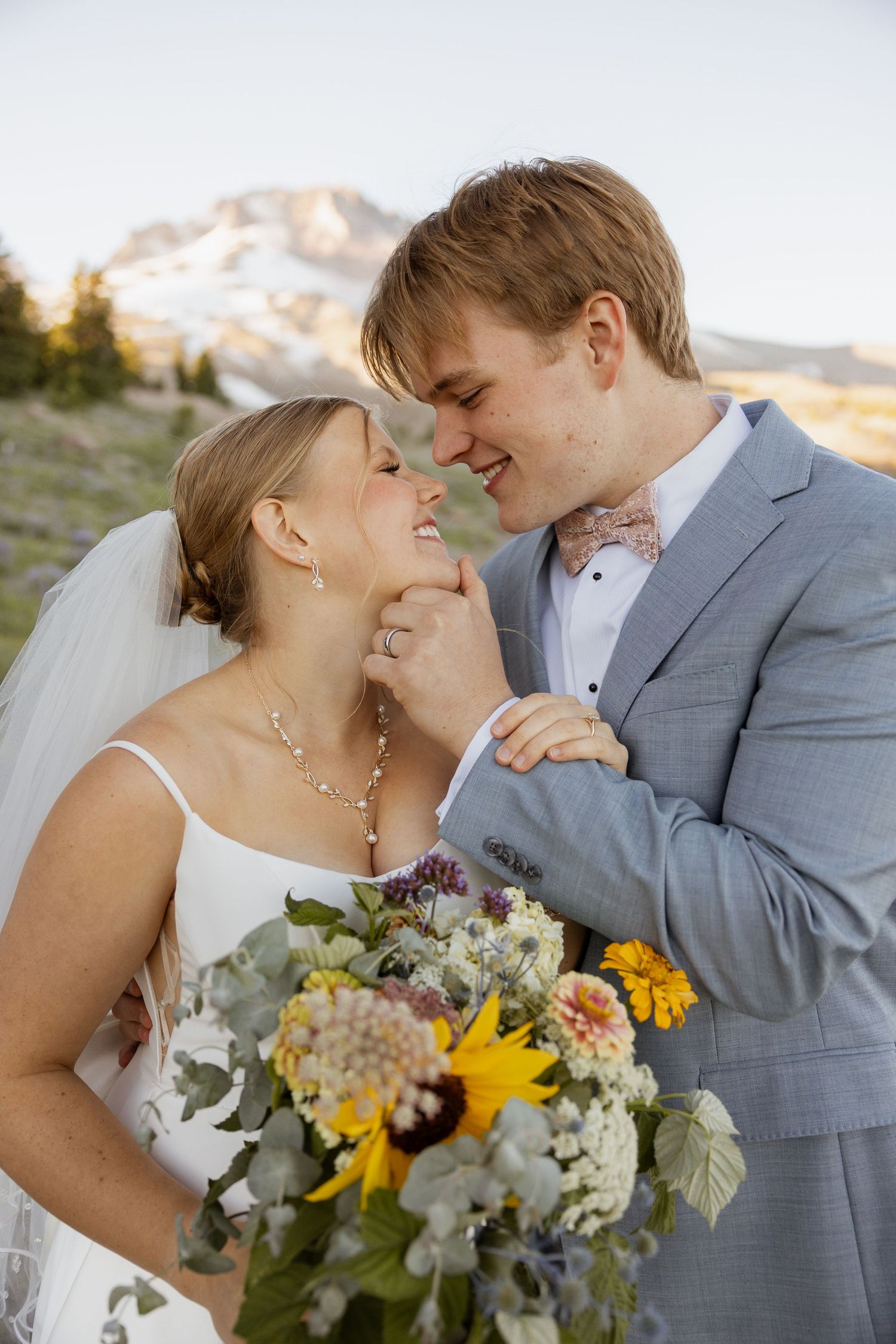 Romantic wedding couple embracing outdoors with sunflowers and mountain scenery.