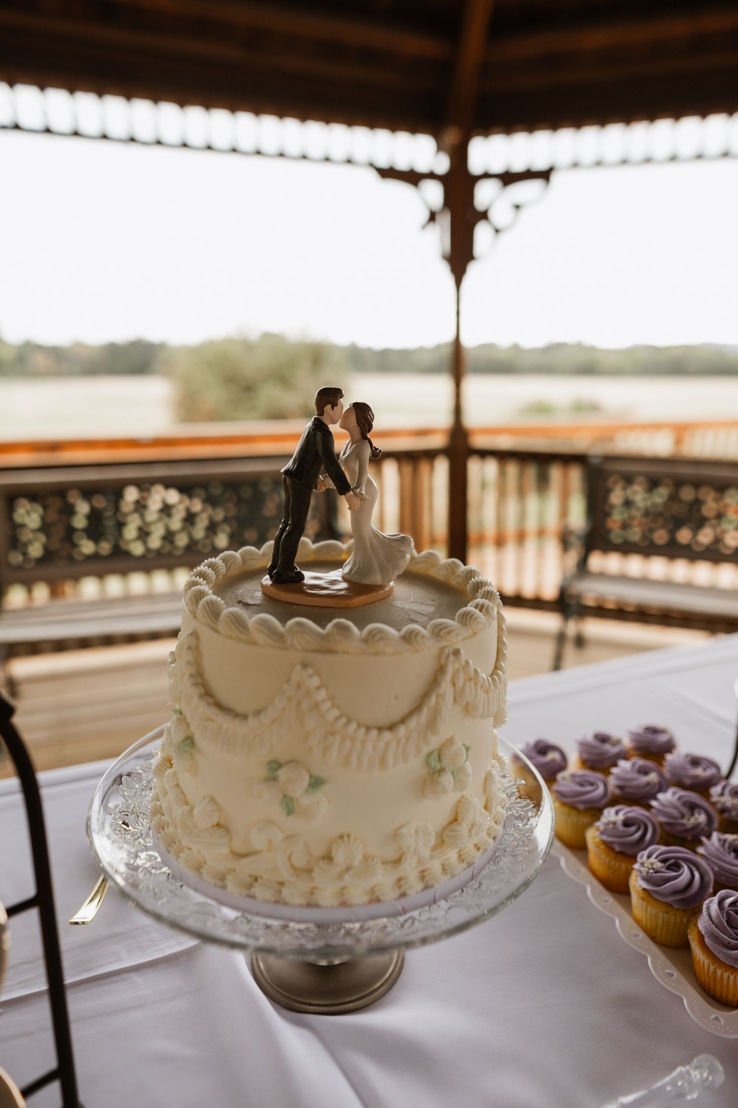 White wedding cake with cupcakes displayed on a covered outdoor deck.