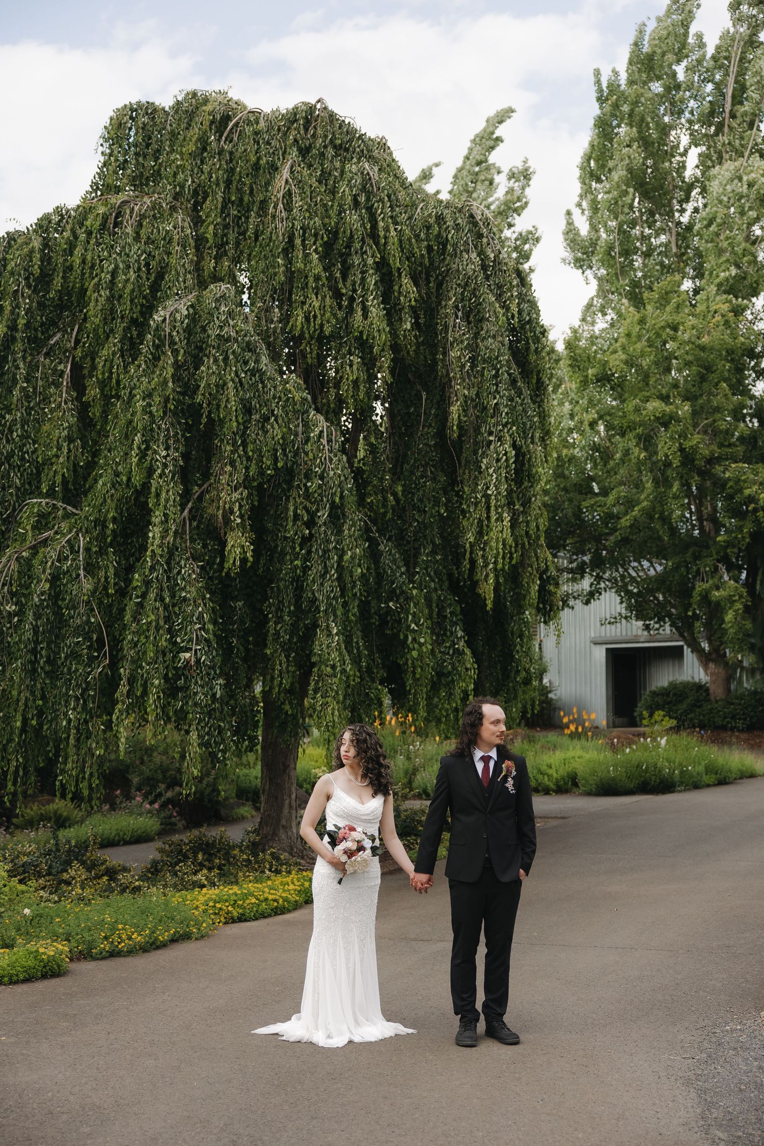 Bride and groom hold hands on a path beneath a weeping willow tree at an outdoor wedding venue.