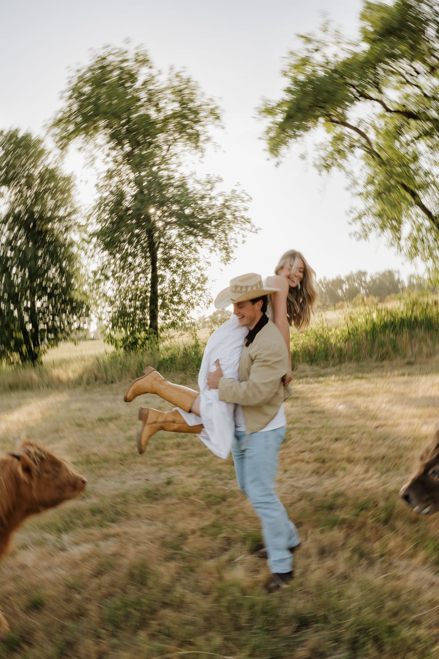 A man in a cowboy hat carries a woman through a sunny rural field with cattle and green trees in the background.
