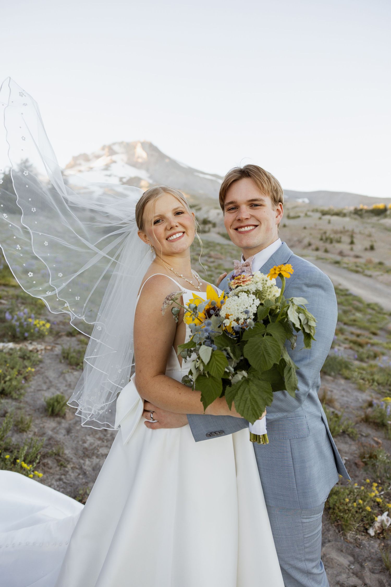 Newlyweds share joyful moments together on a mountainside with sunflowers and a flowing white veil.