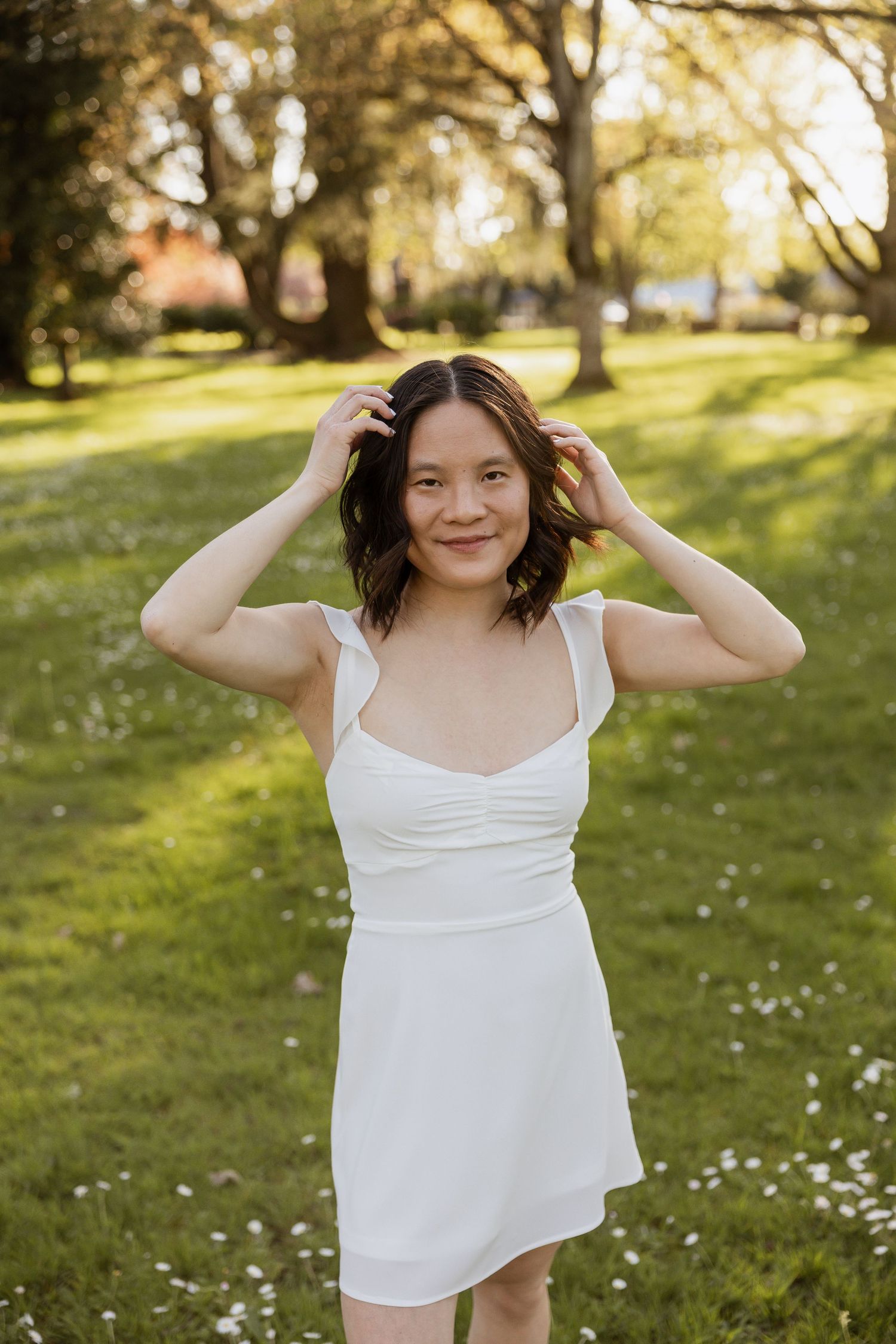 A person in a white sundress poses in a sunny park with golden sunlight filtering through trees in the background.