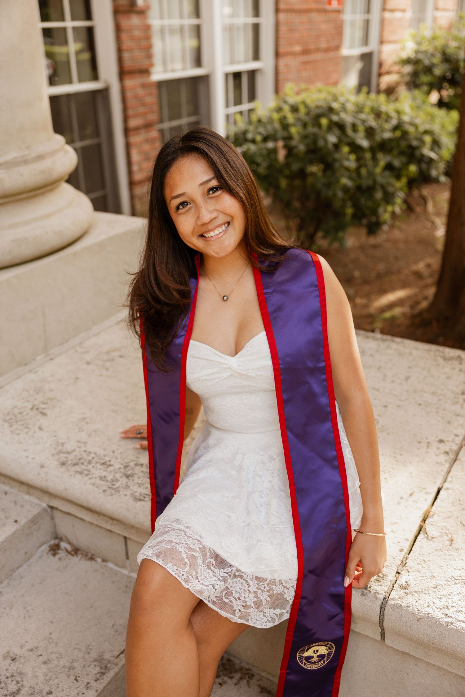 A graduate in a white lace top and navy blue graduation stole poses on stone steps for a series of photos.