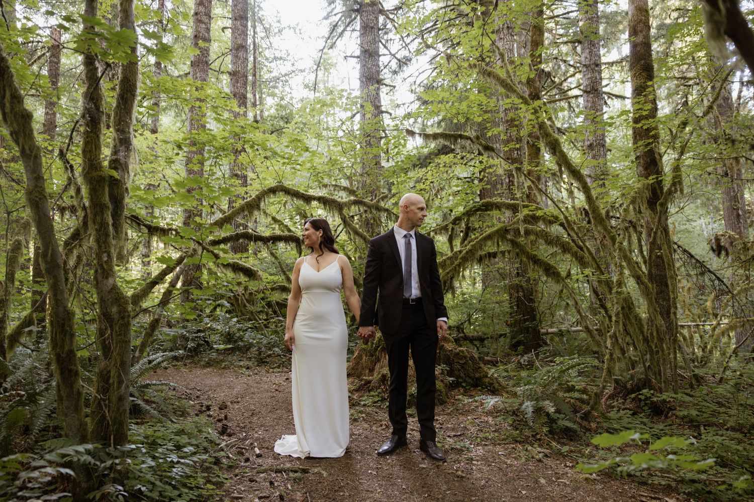 A couple stands together on a forest path surrounded by lush green trees and ferns during a wedding ceremony.