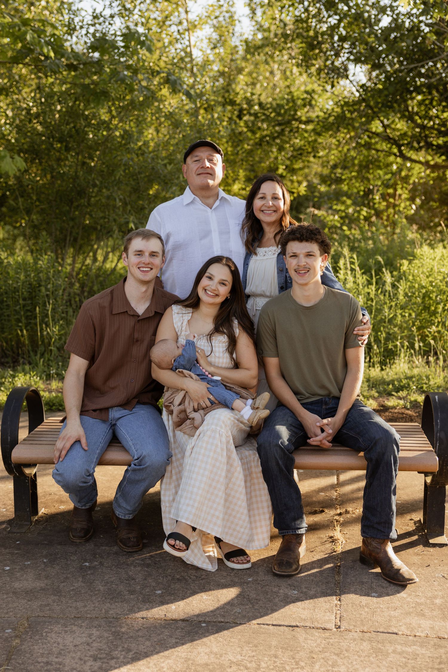 A family sits together on a wooden bench in a sunlit outdoor setting surrounded by green foliage.