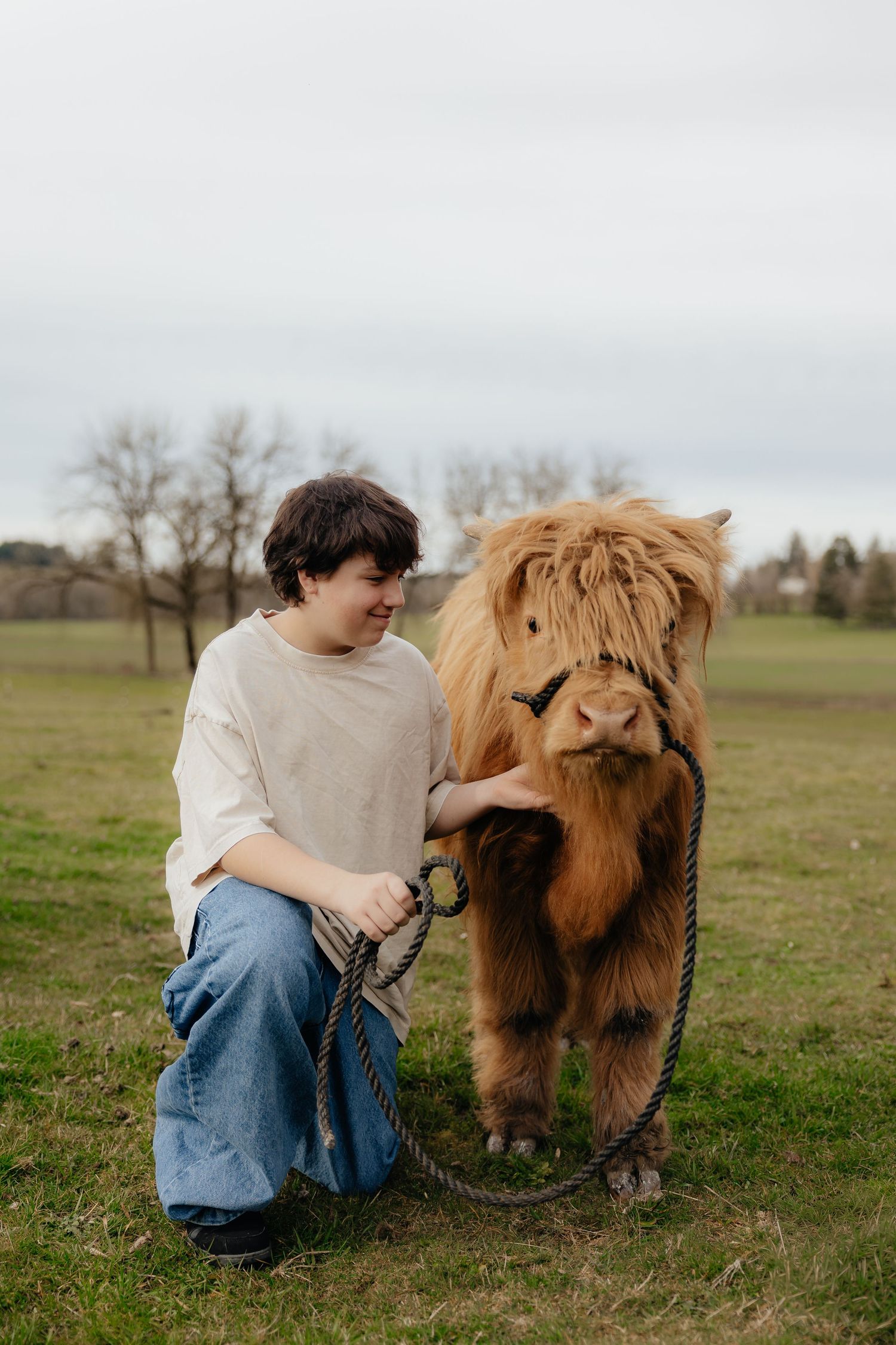A young person kneeling beside a fluffy Highland cow on a green field, gently holding its rope lead.