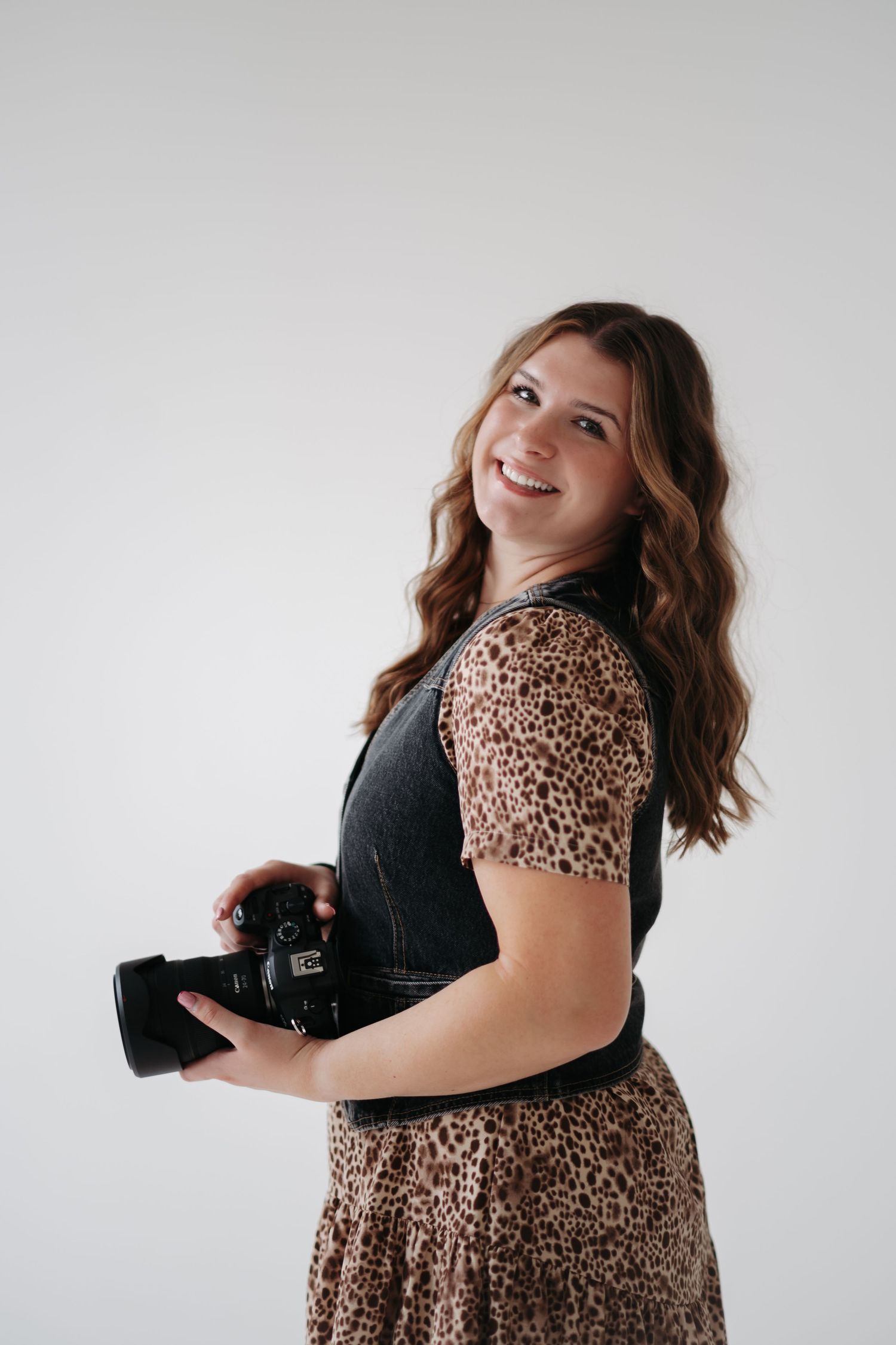 Photographer in animal print dress and black vest poses playfully with camera in studio setting.