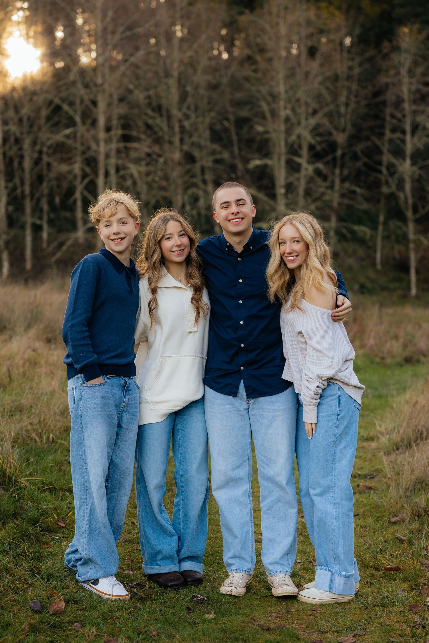 Four young people in coordinated denim and blue outfits pose together outdoors at sunset in front of bare trees.