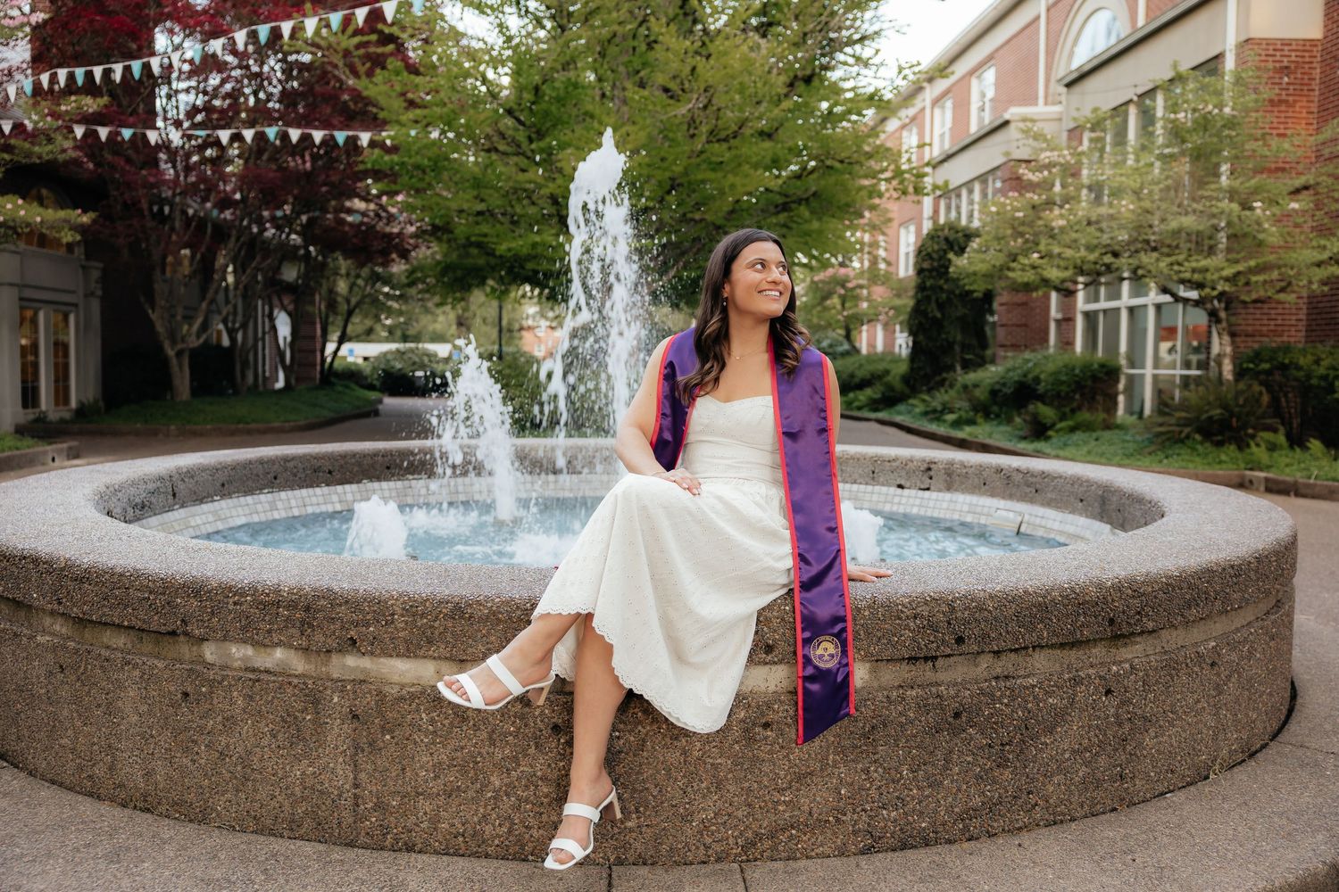 Graduate in white dress and purple stole sits by a fountain on a college campus on a sunny day.