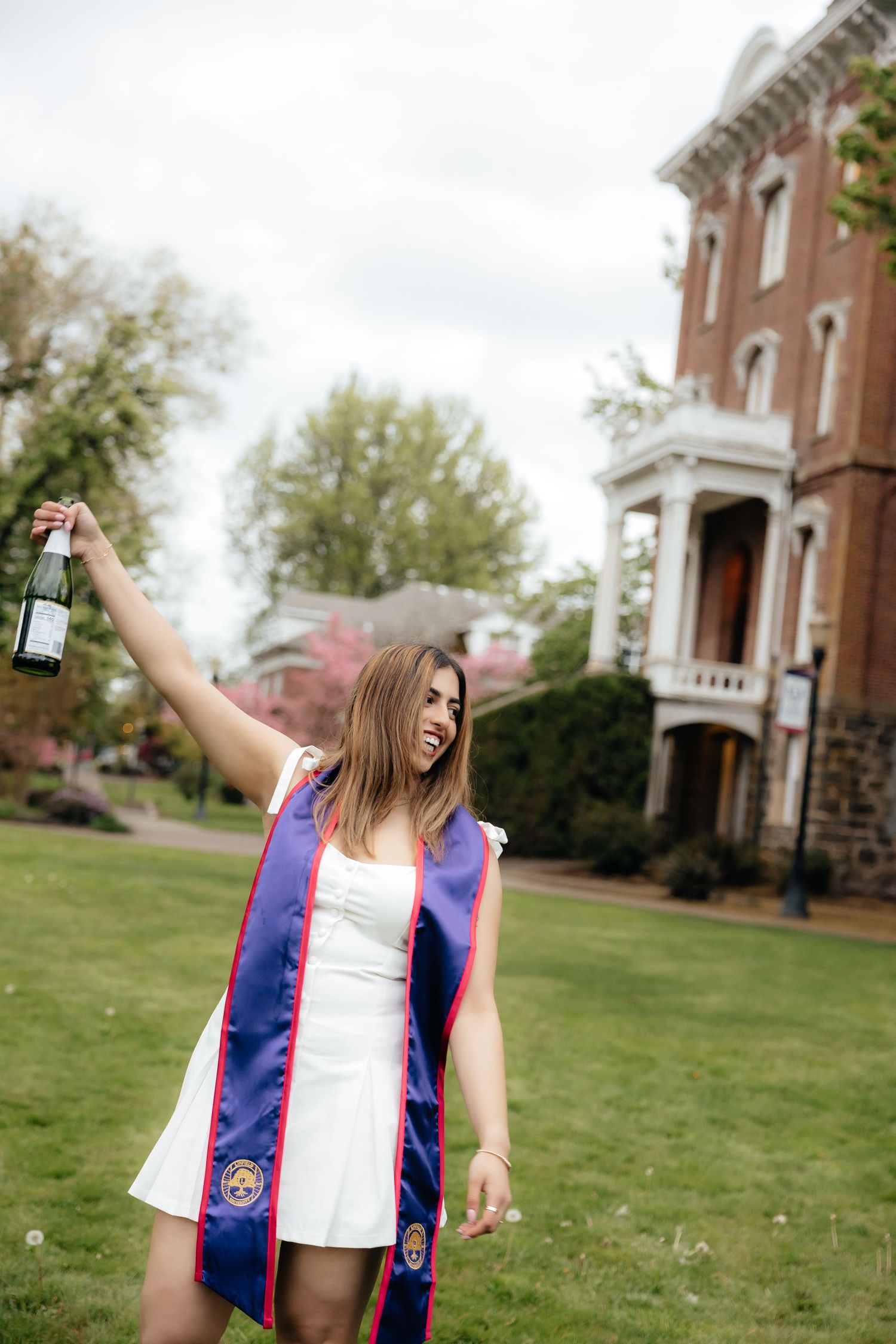 Young woman in white dress with graduation sash celebrates outdoors, raising a bottle near a brick building.