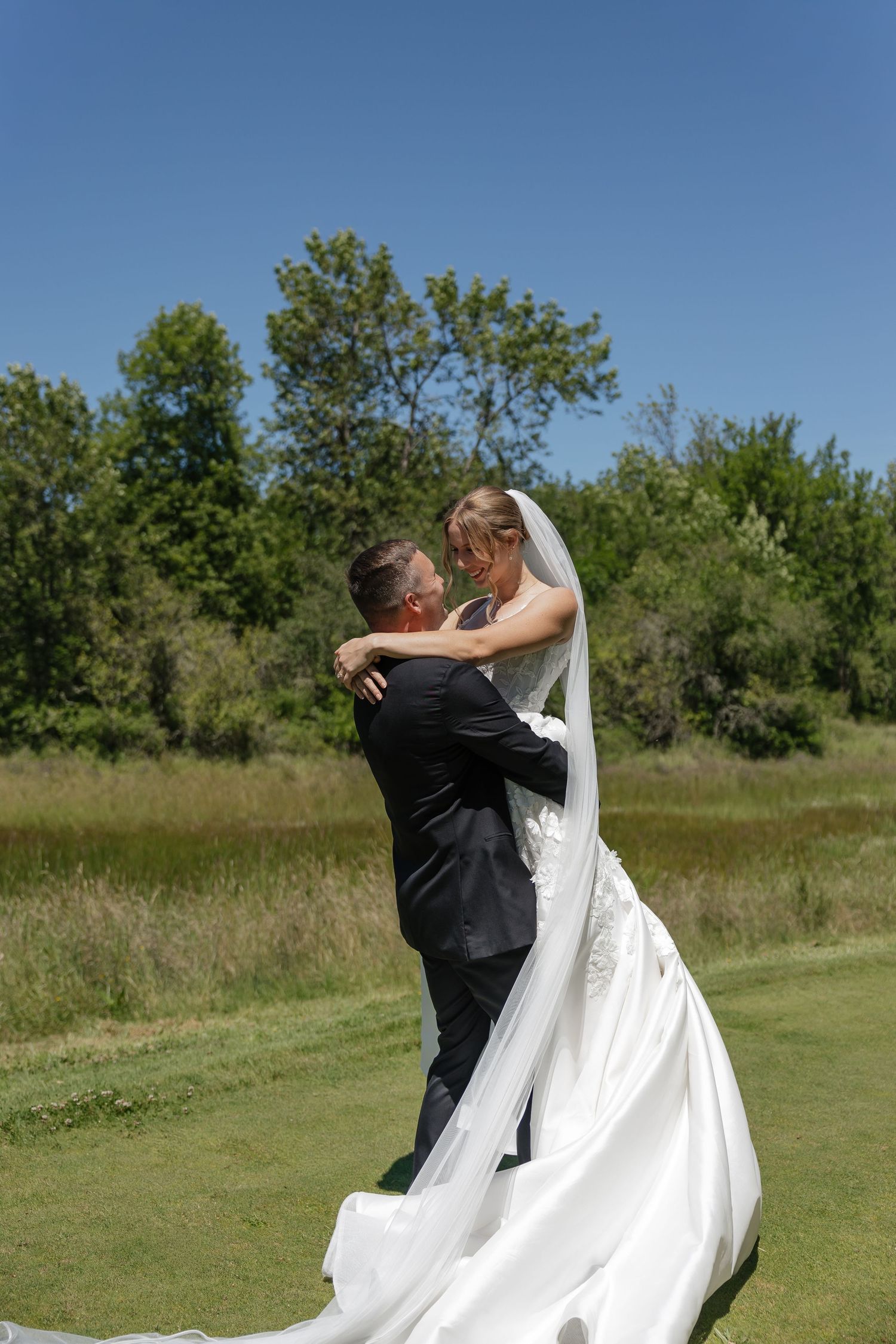 Bride and groom embrace on green lawn, her white gown and veil flowing, surrounded by lush trees under a clear blue sky.