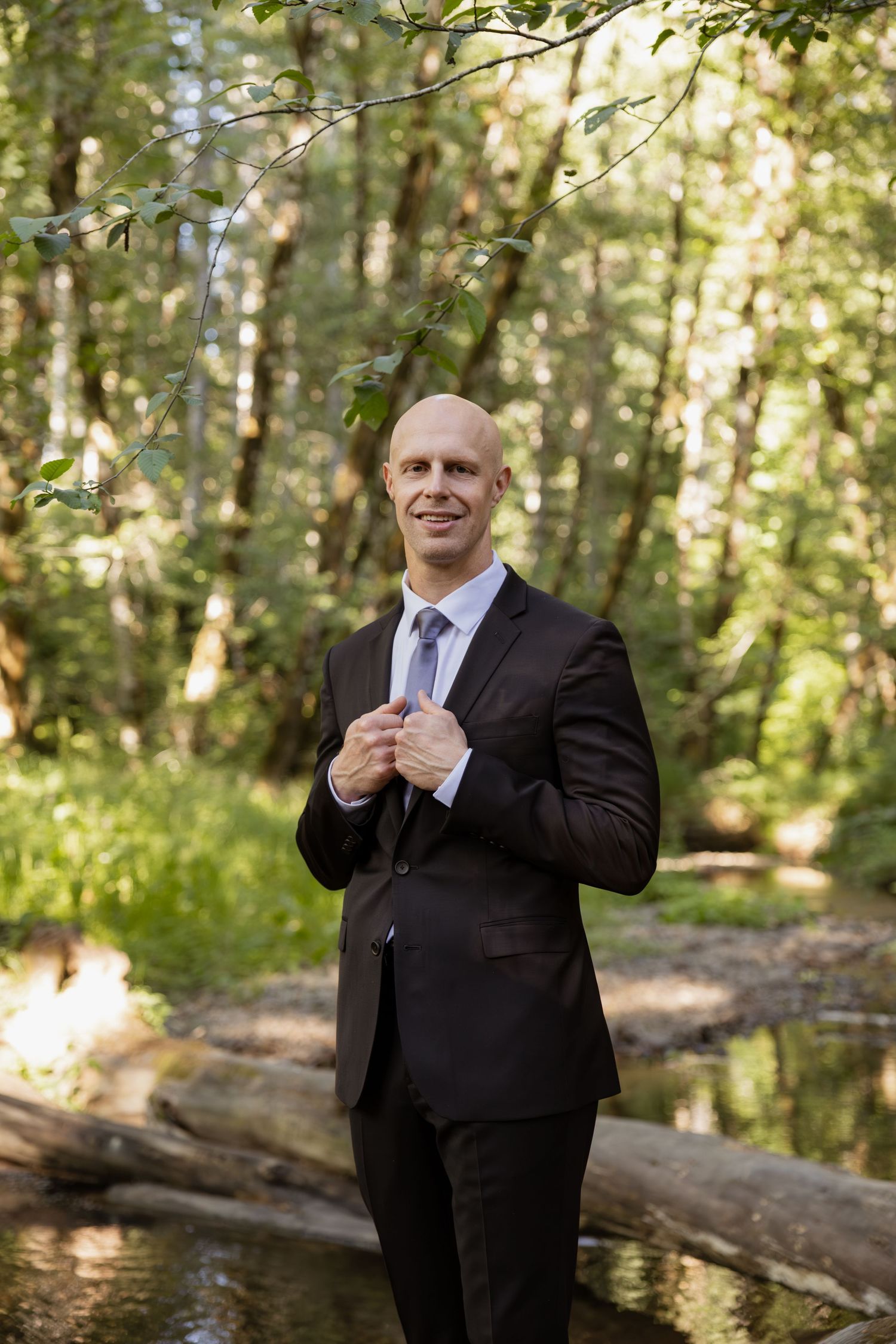 A person in a black suit gestures playfully while standing in a sunlit forest setting.