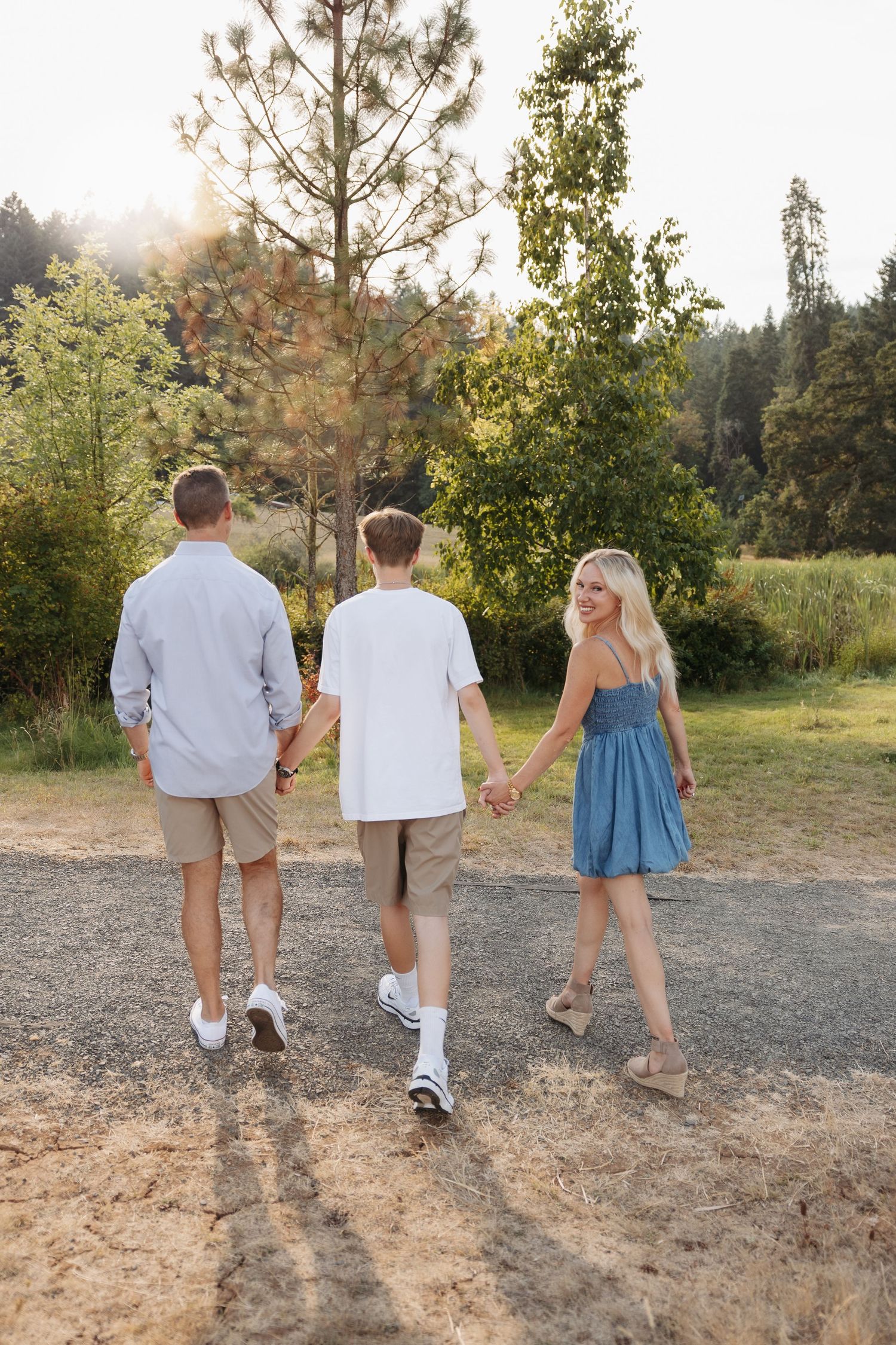 Three people walking hand in hand on a dirt path surrounded by trees and greenery in warm afternoon sunlight.