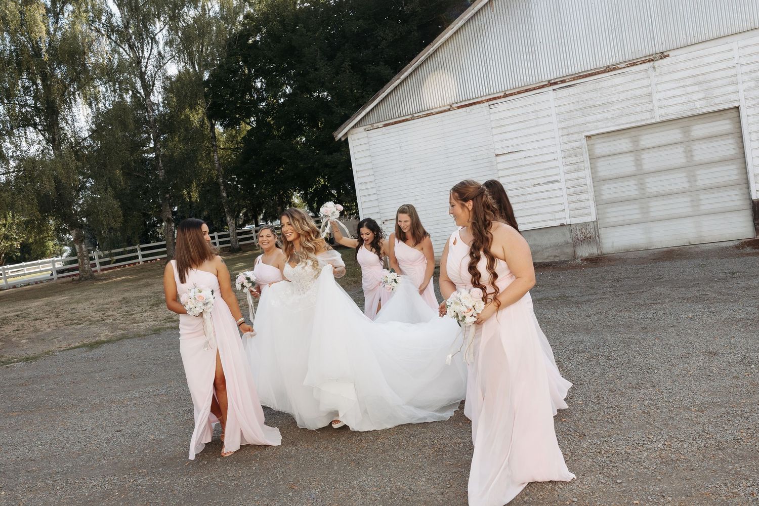 Bridesmaids in blush pink dresses walking near white barn on wedding day.