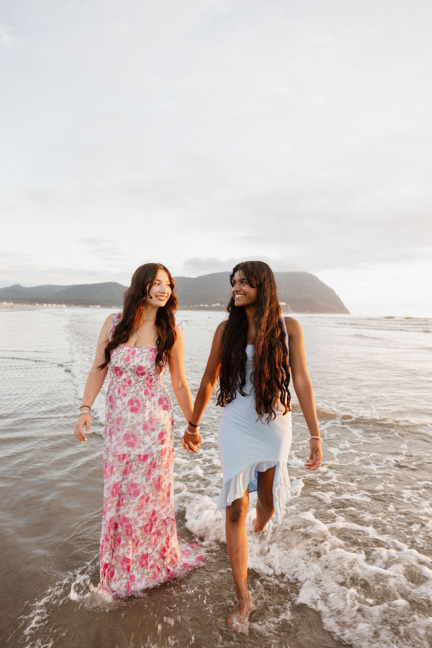 Two friends in summer dresses laugh and splash together in ocean waves at sunset along a scenic beach.