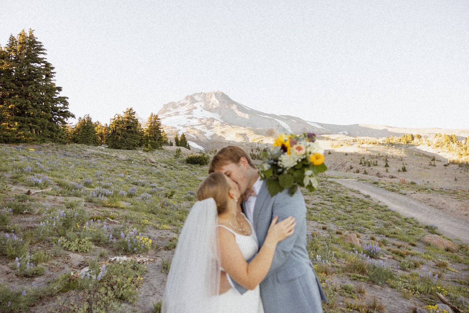 A couple shares a romantic moment on a mountainside with Mount Hood visible in the background at sunset.