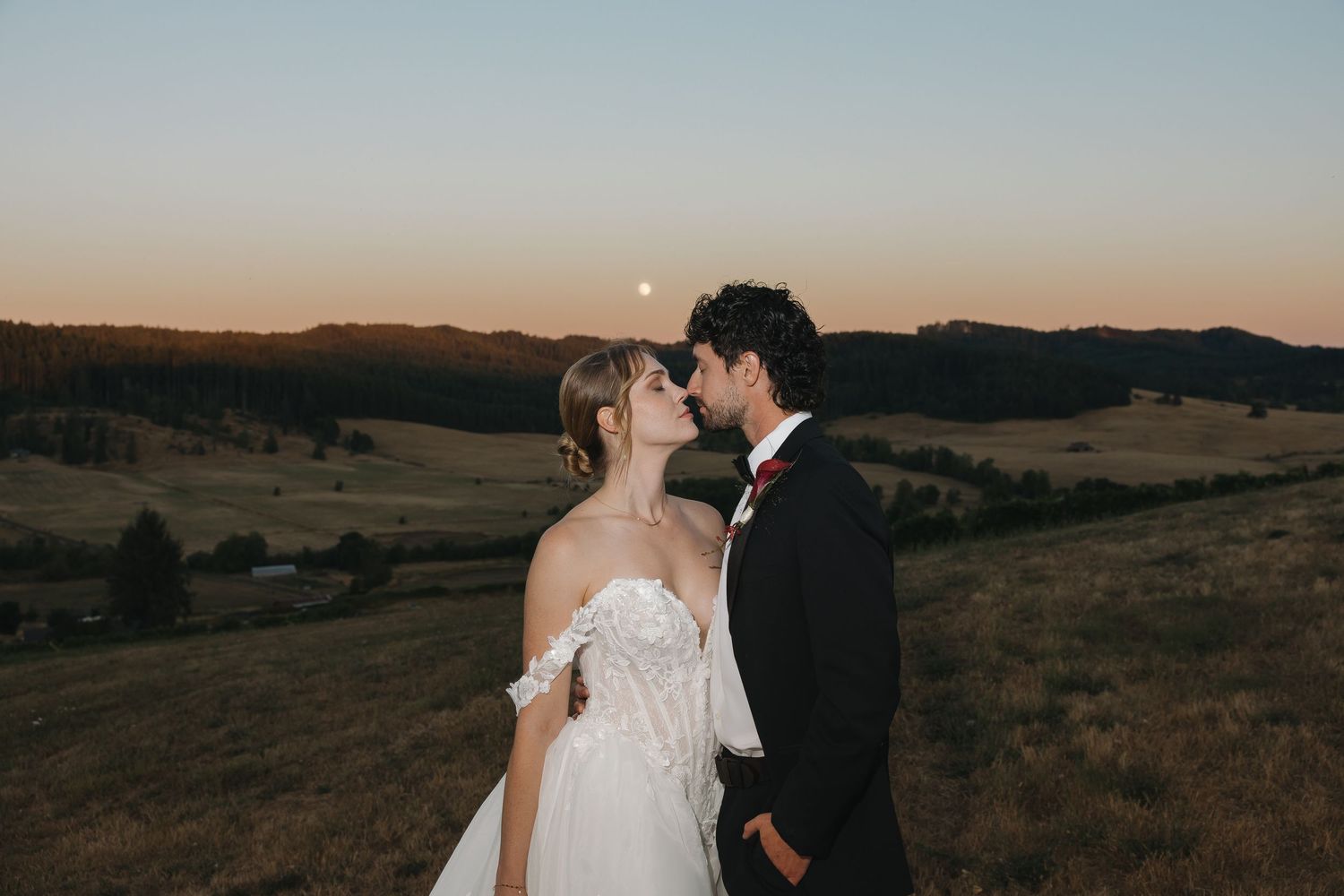 A bride and groom share a romantic kiss at sunset on a scenic hilltop with rolling green fields and a full moon rising.