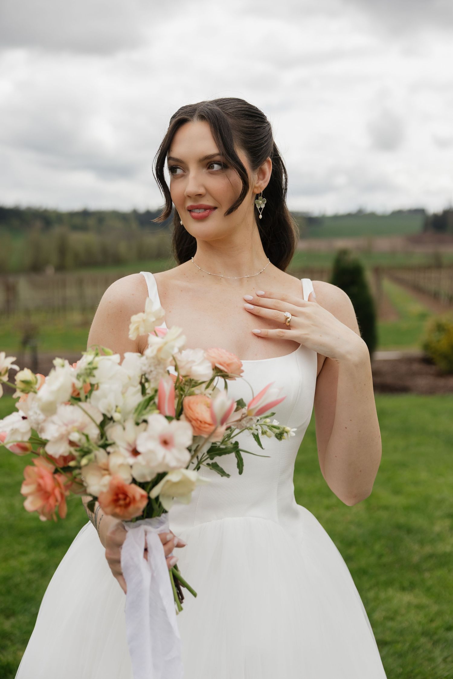 Bride in white gown holding peach and white floral bouquet, posing outdoors at vineyard wedding venue.
