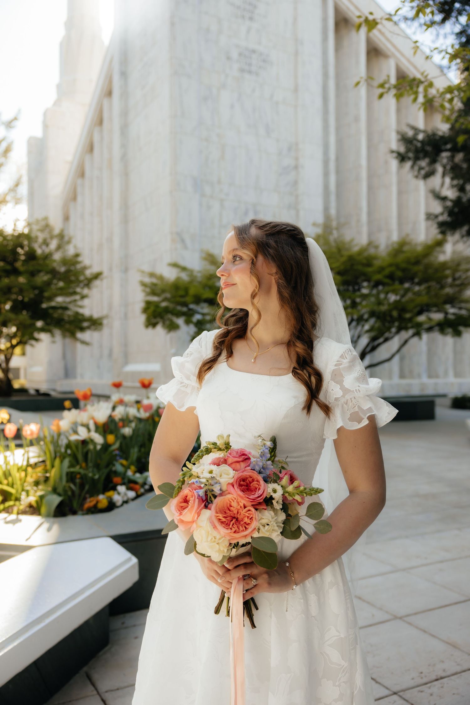 Bride in white lace dress with veil holds colorful bouquet outside marble temple building.