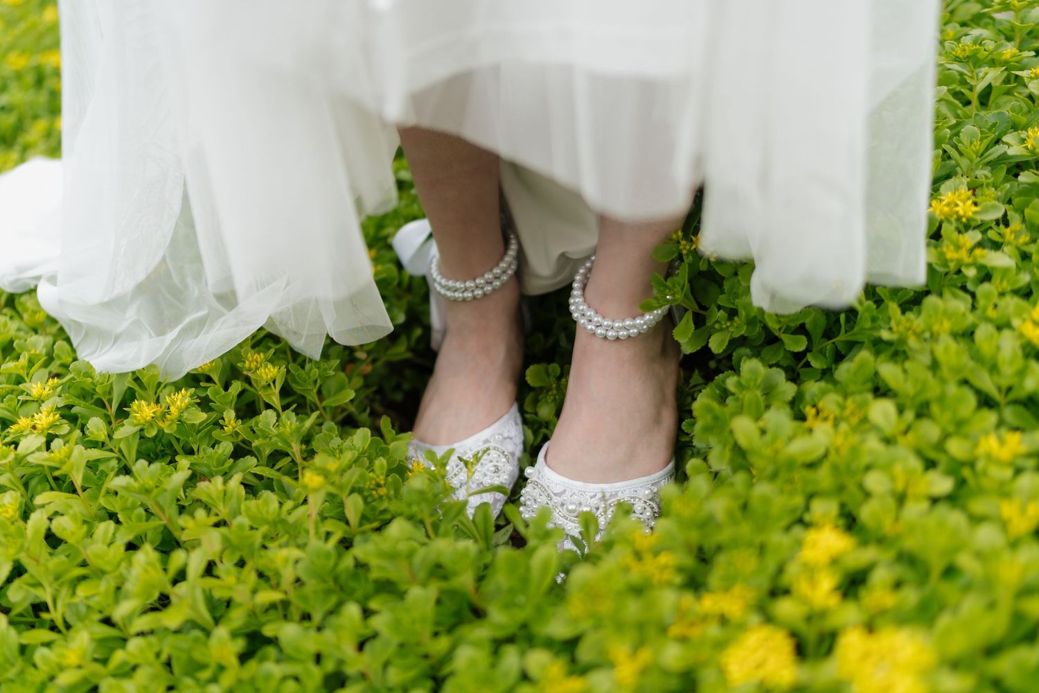 Bride wearing white lace flats with pearl ankle straps standing on lush green ground cover with yellow flowers.