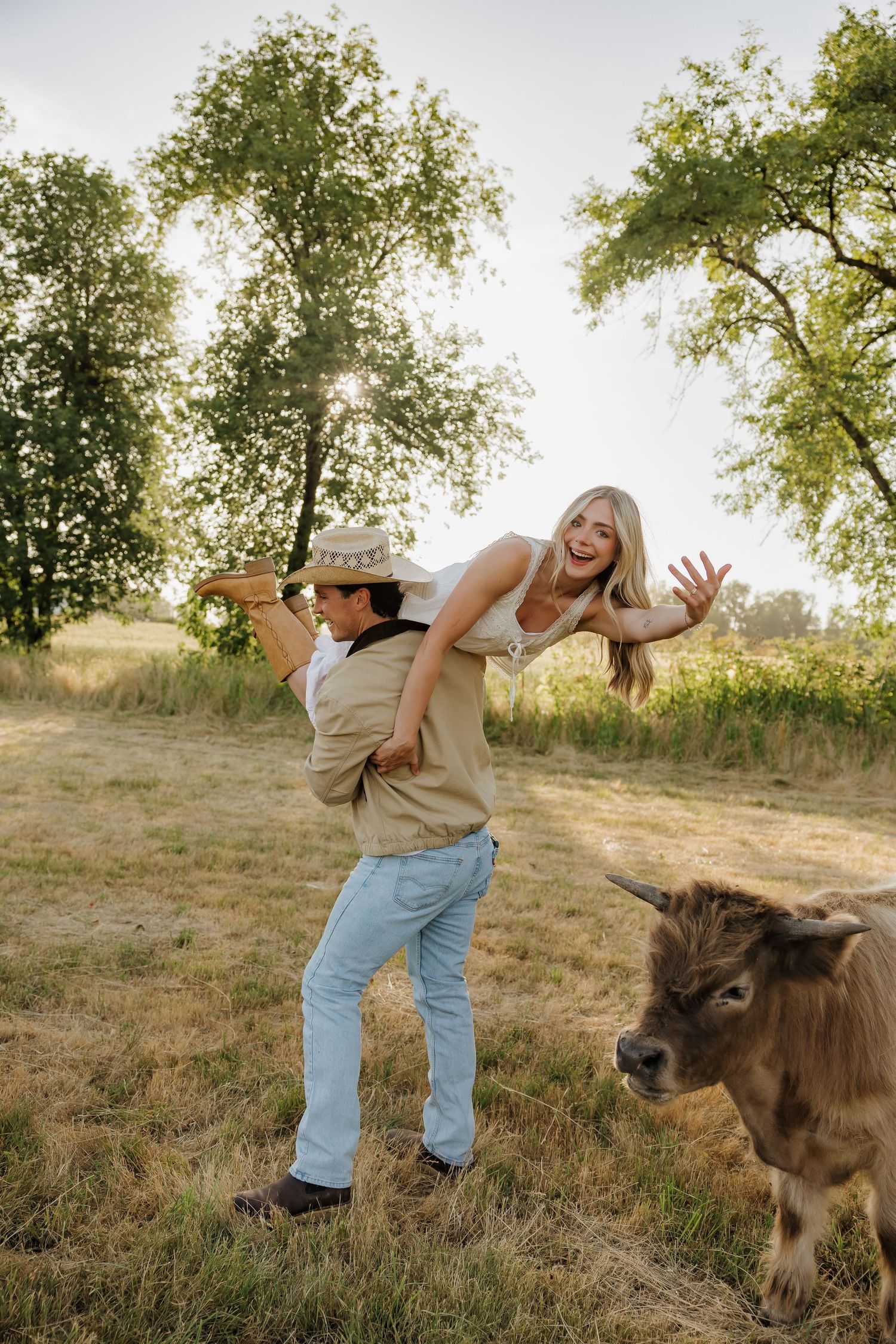 A joyful couple plays in a sunny field, man in cowboy hat carrying laughing woman over shoulder near a small brown bull.