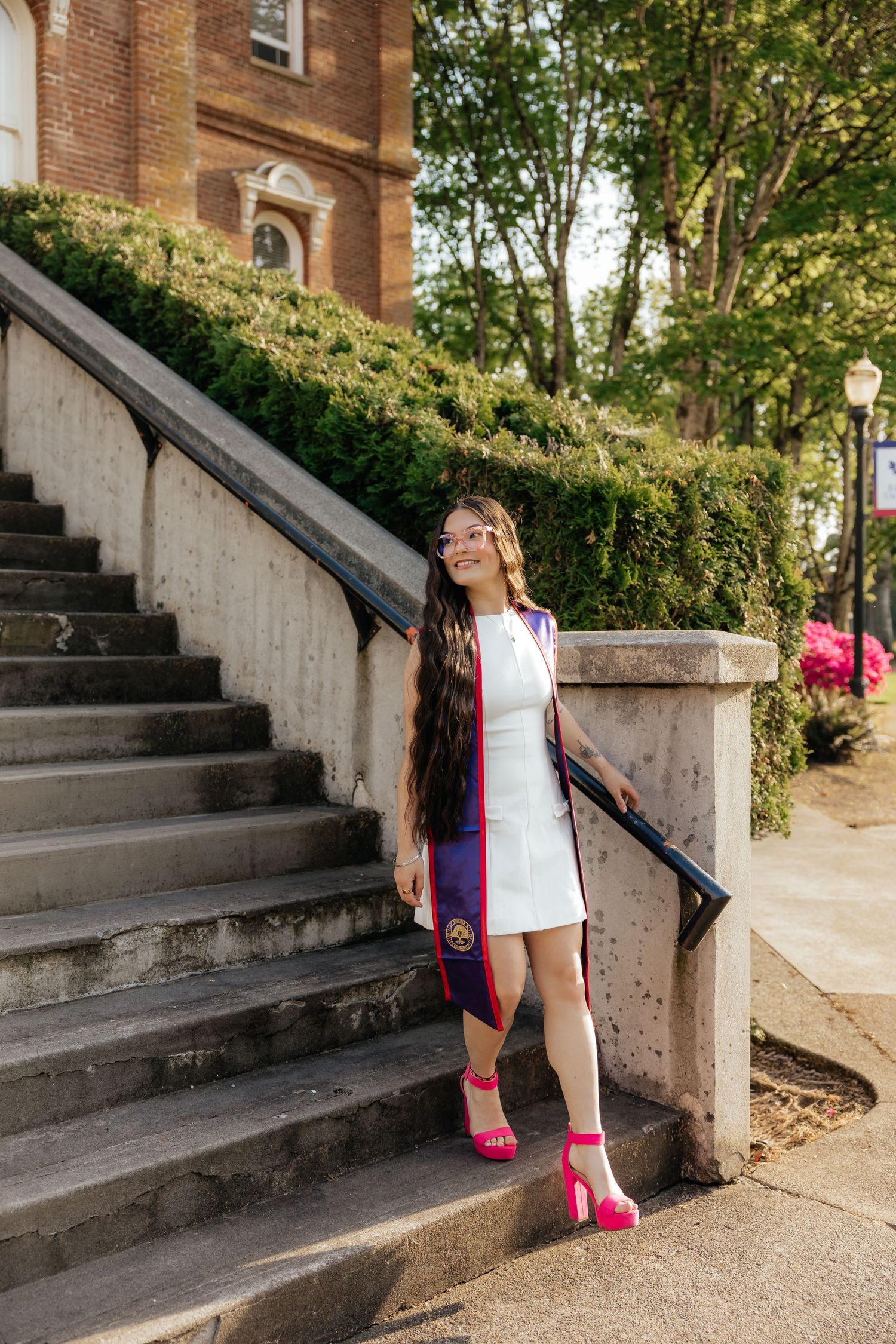 Young woman in white dress and graduation stole poses on stone steps outside a brick university building.
