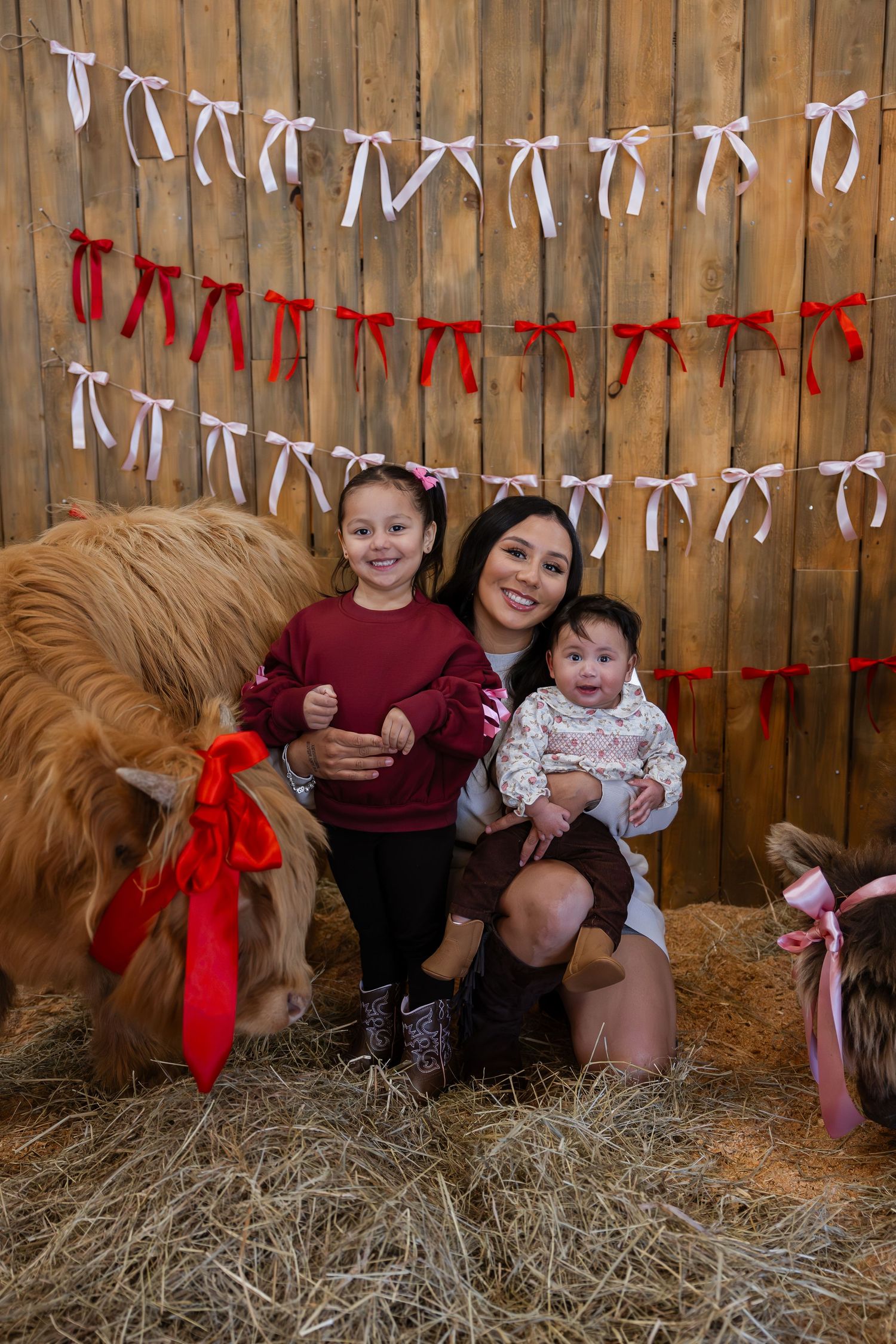 A group sits on hay bales in front of a wooden wall decorated with red and white ribbon garlands for holiday photos.
