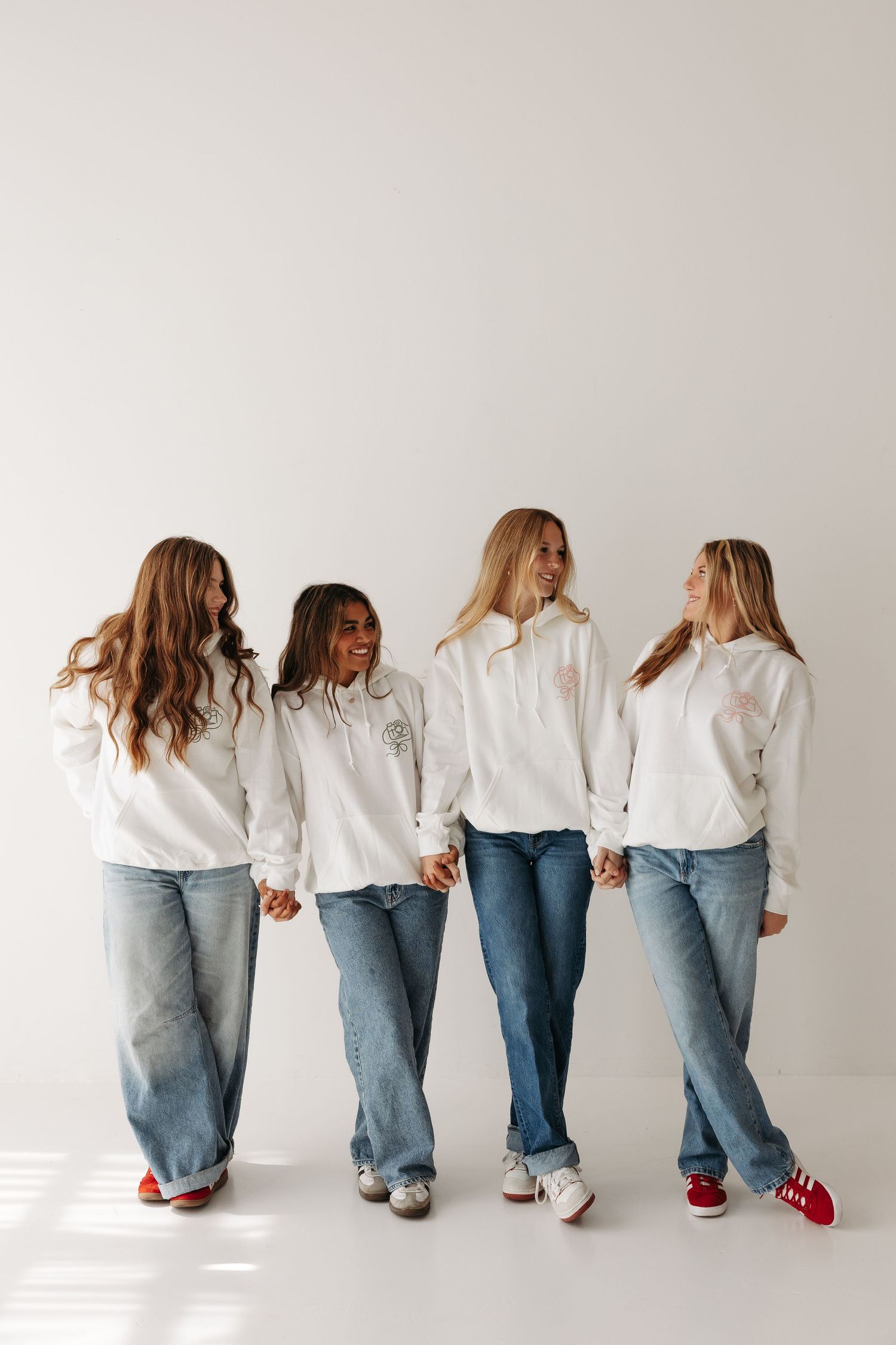 Four young women in matching white hoodies and blue jeans hold hands, smiling together against a white background.