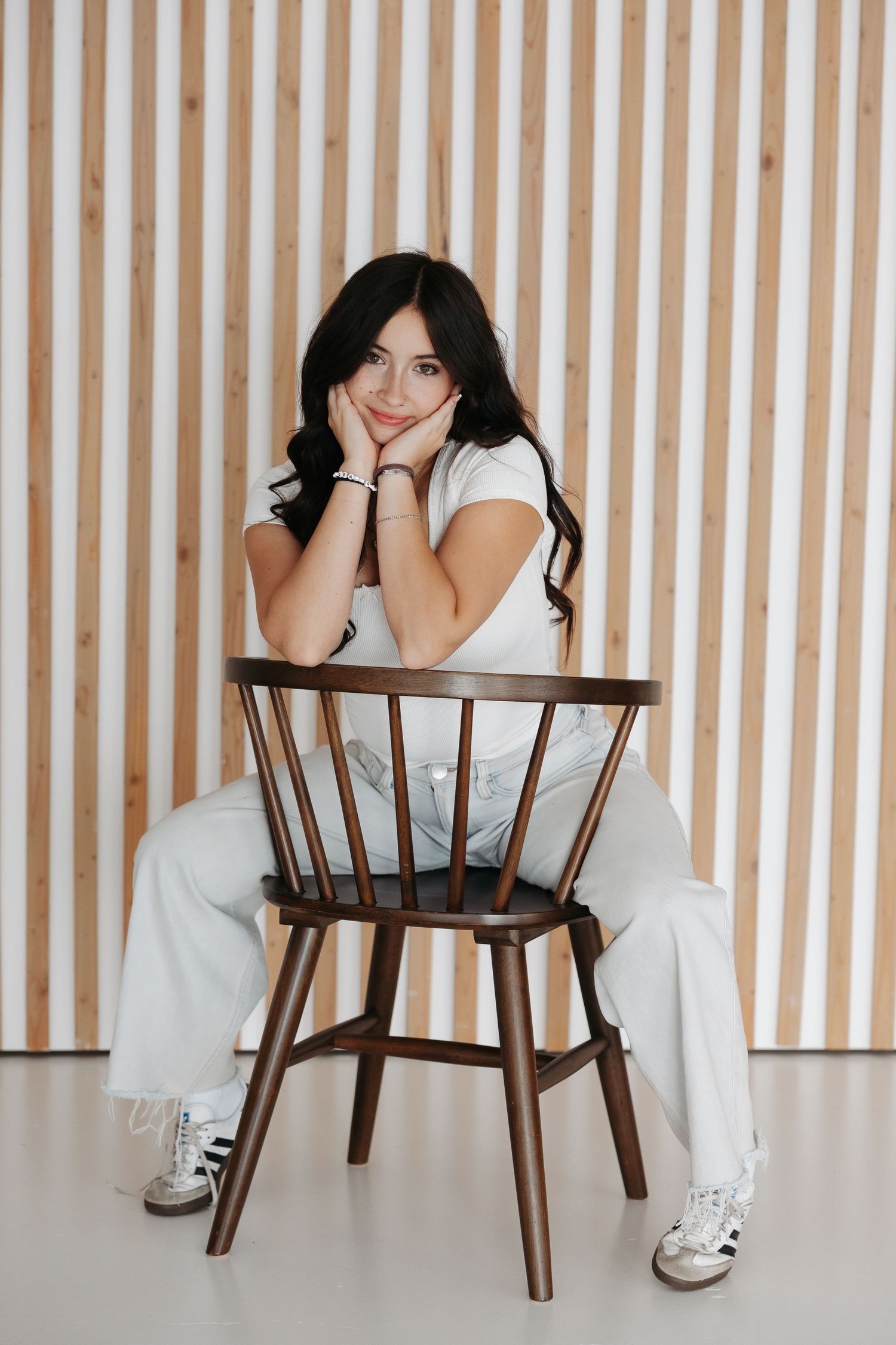 Person in white dress sits on wooden chair against striped wall background in minimalist photo series.