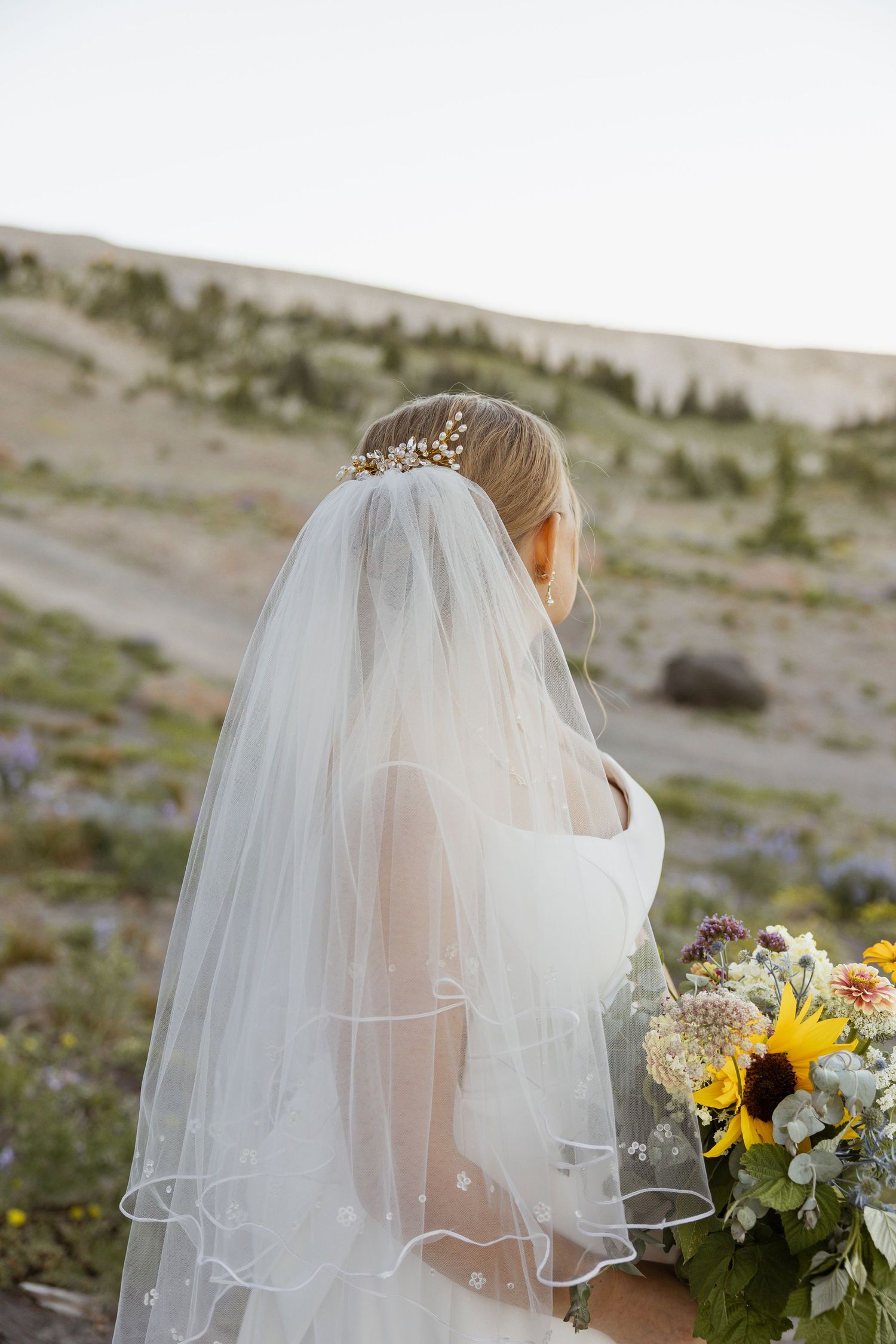 A bride in a flowing white dress and veil holds a sunflower bouquet against a mountain landscape.