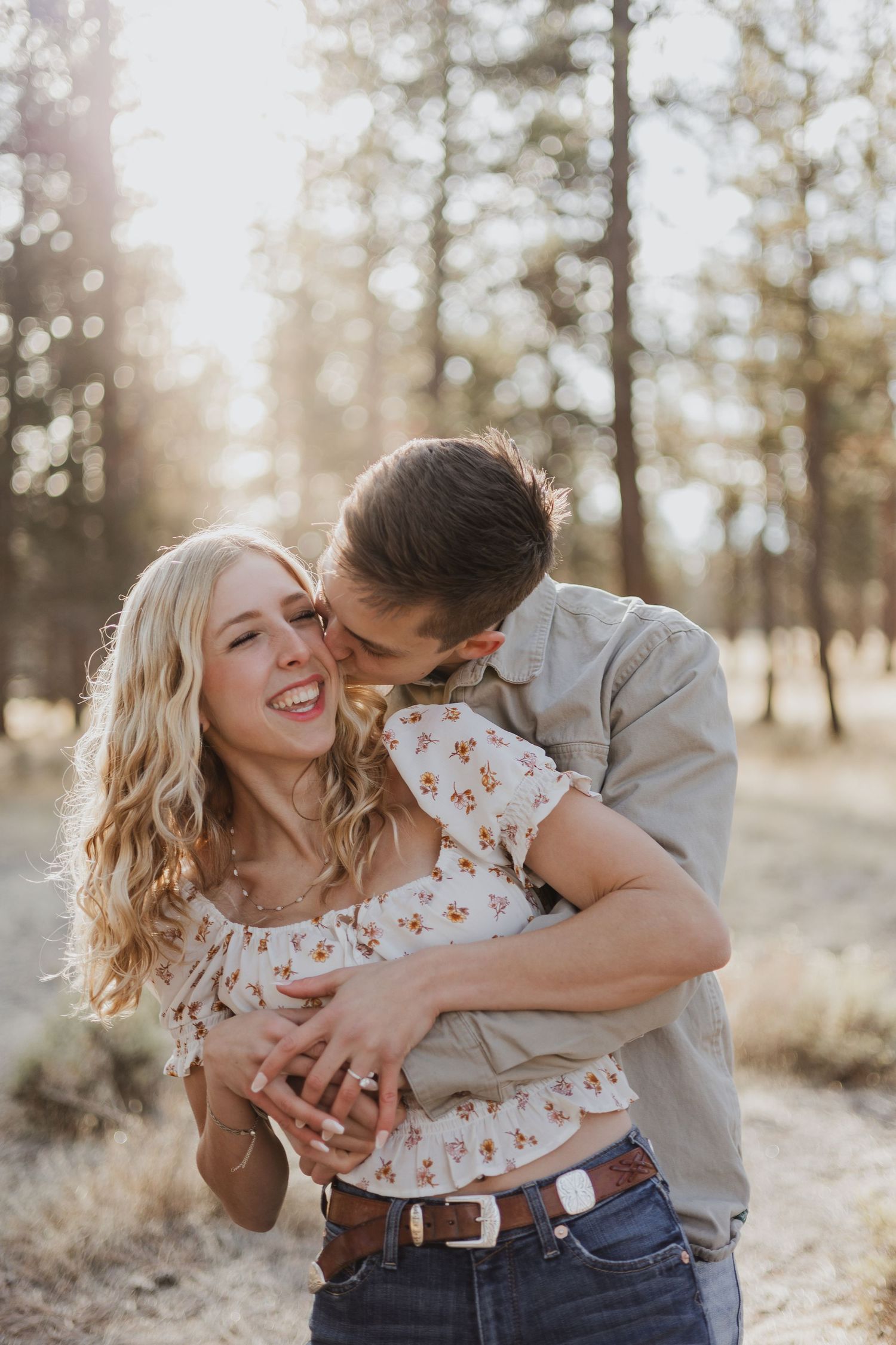Playful couple embracing and laughing in golden sunlit forest setting.