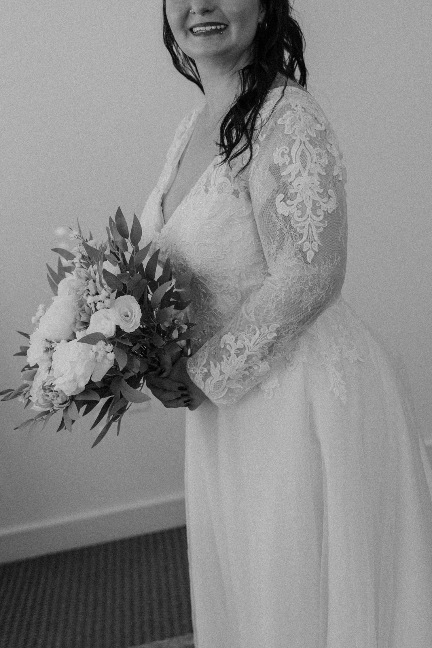 A bride in a long-sleeved lace wedding gown holds an elegant floral bouquet in a black and white photograph.