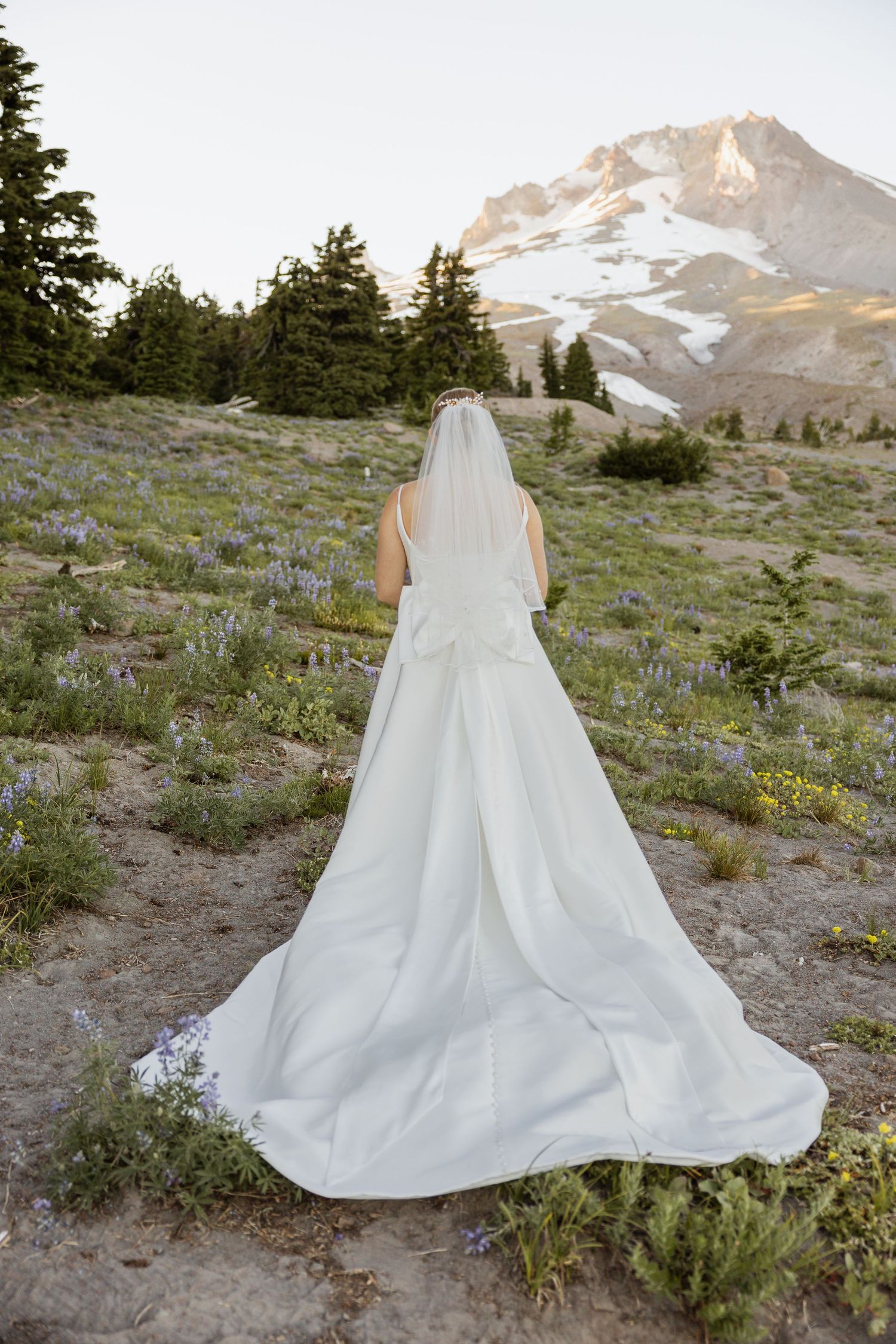 A flowing white wedding dress displayed on a mountain slope with wildflowers and Mount Hood in the background.