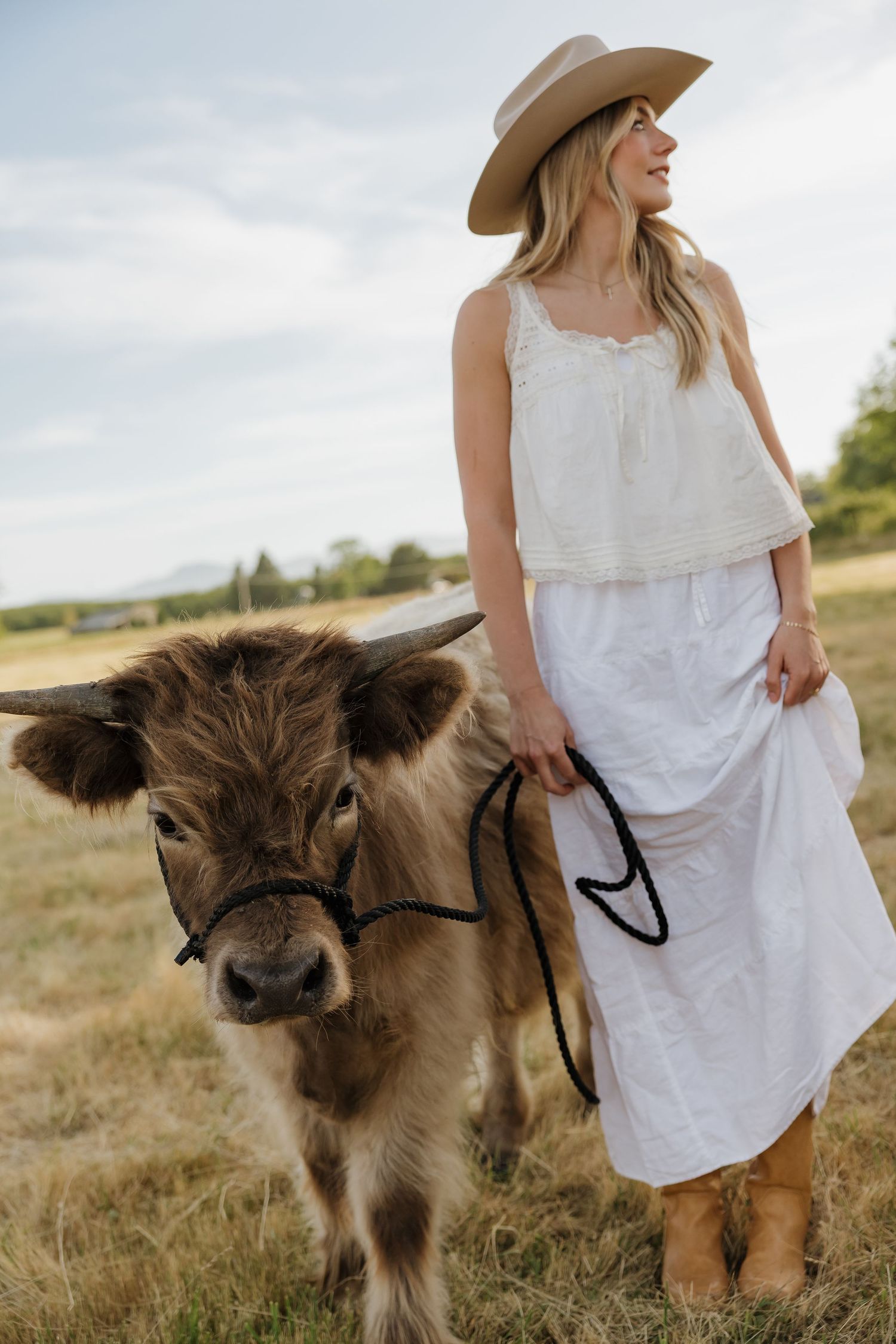 Smiling woman in white dress and cowboy hat leading a Highland calf on a leash in a sunny field.