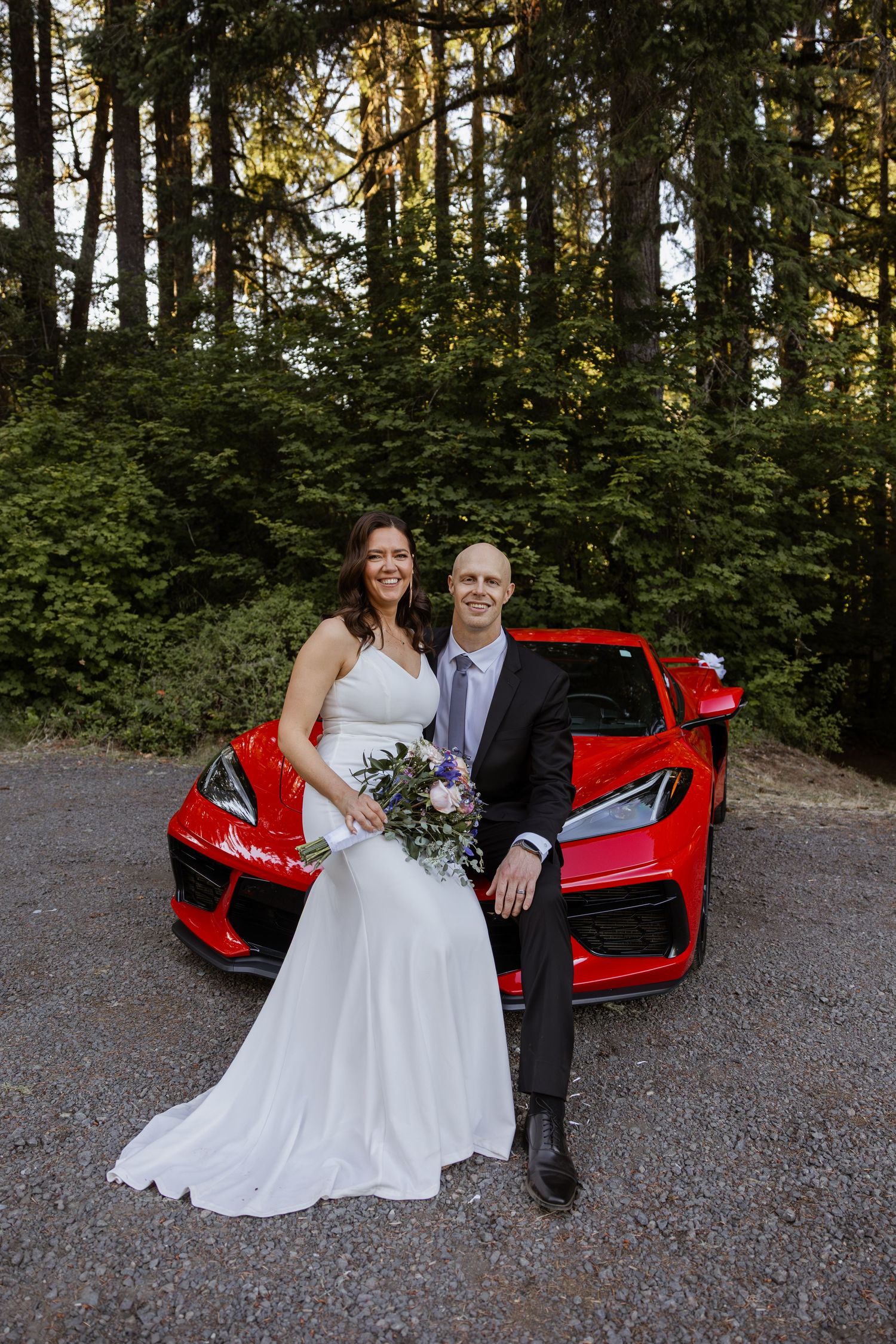 A couple poses next to a bright red sports car in a forest setting during their wedding photoshoot.