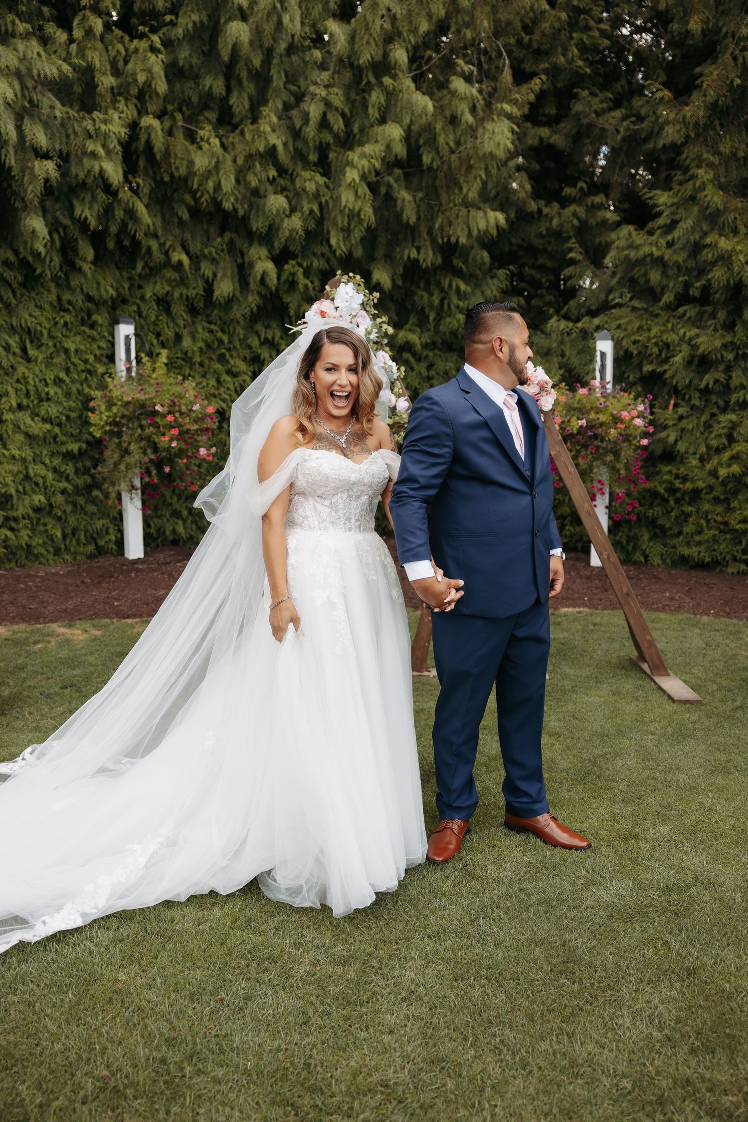 A couple in wedding attire share a romantic moment outdoors on a grassy lawn with trees in the background.
