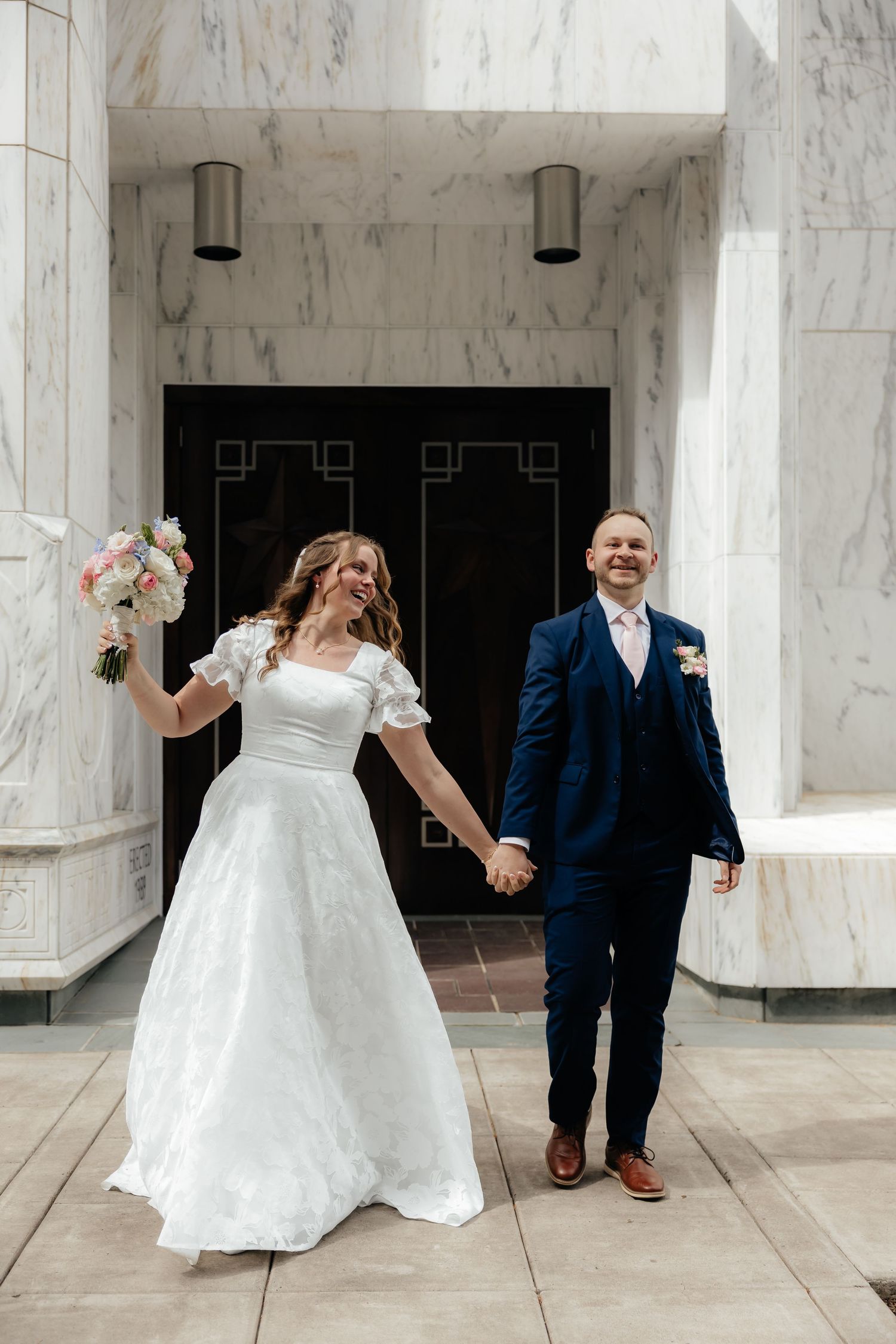 Happy newlyweds hold hands outside marble temple, bride in white dress, groom in navy suit.