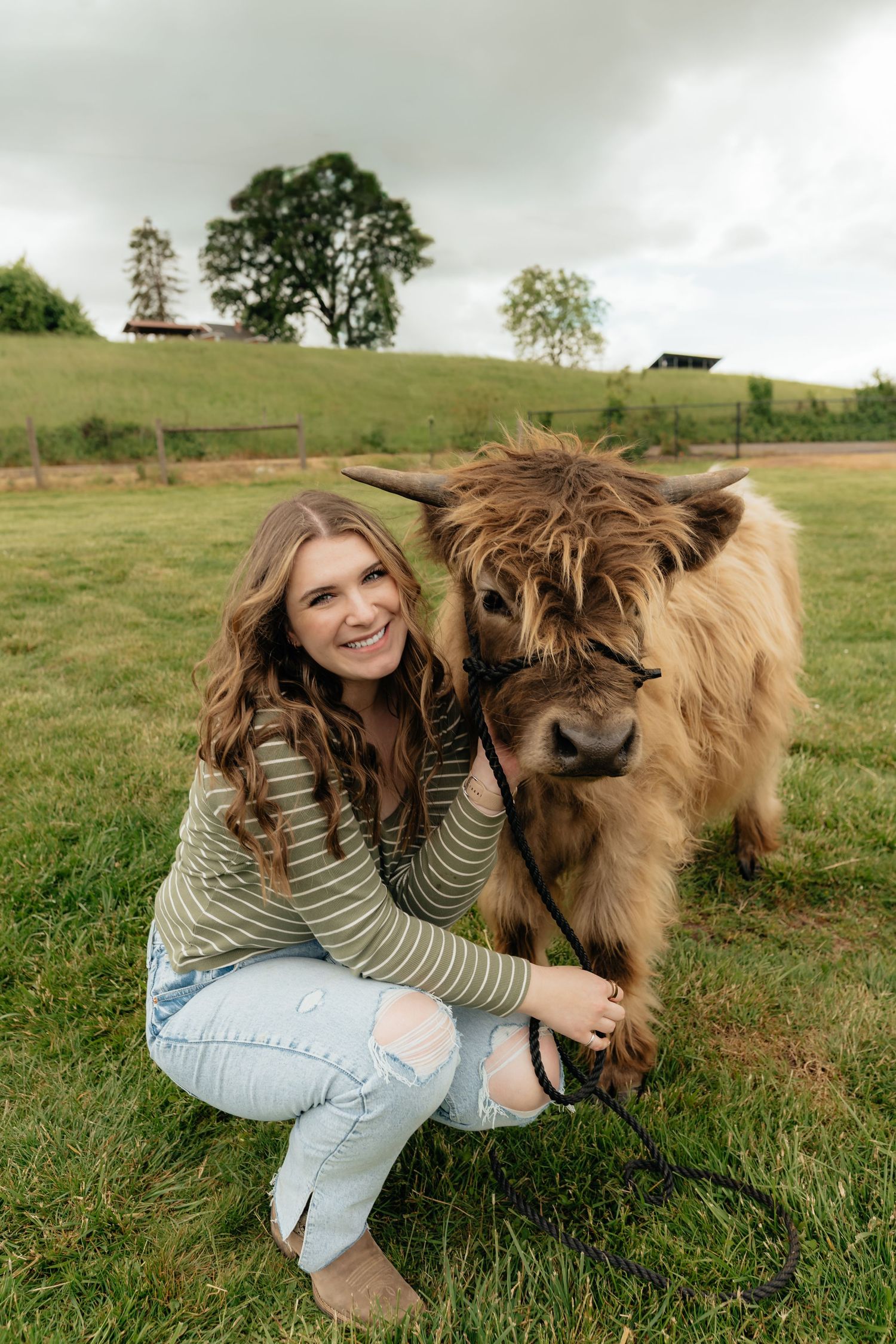 A smiling young woman crouches beside a fluffy Highland cow on a lush green farm pasture.
