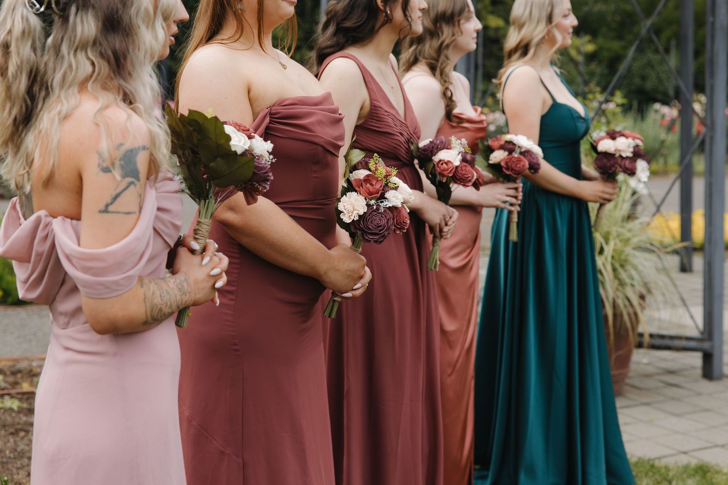 Bridesmaids in mismatched jewel-tone dresses holding bouquets of red and white flowers stand in a row outdoors.