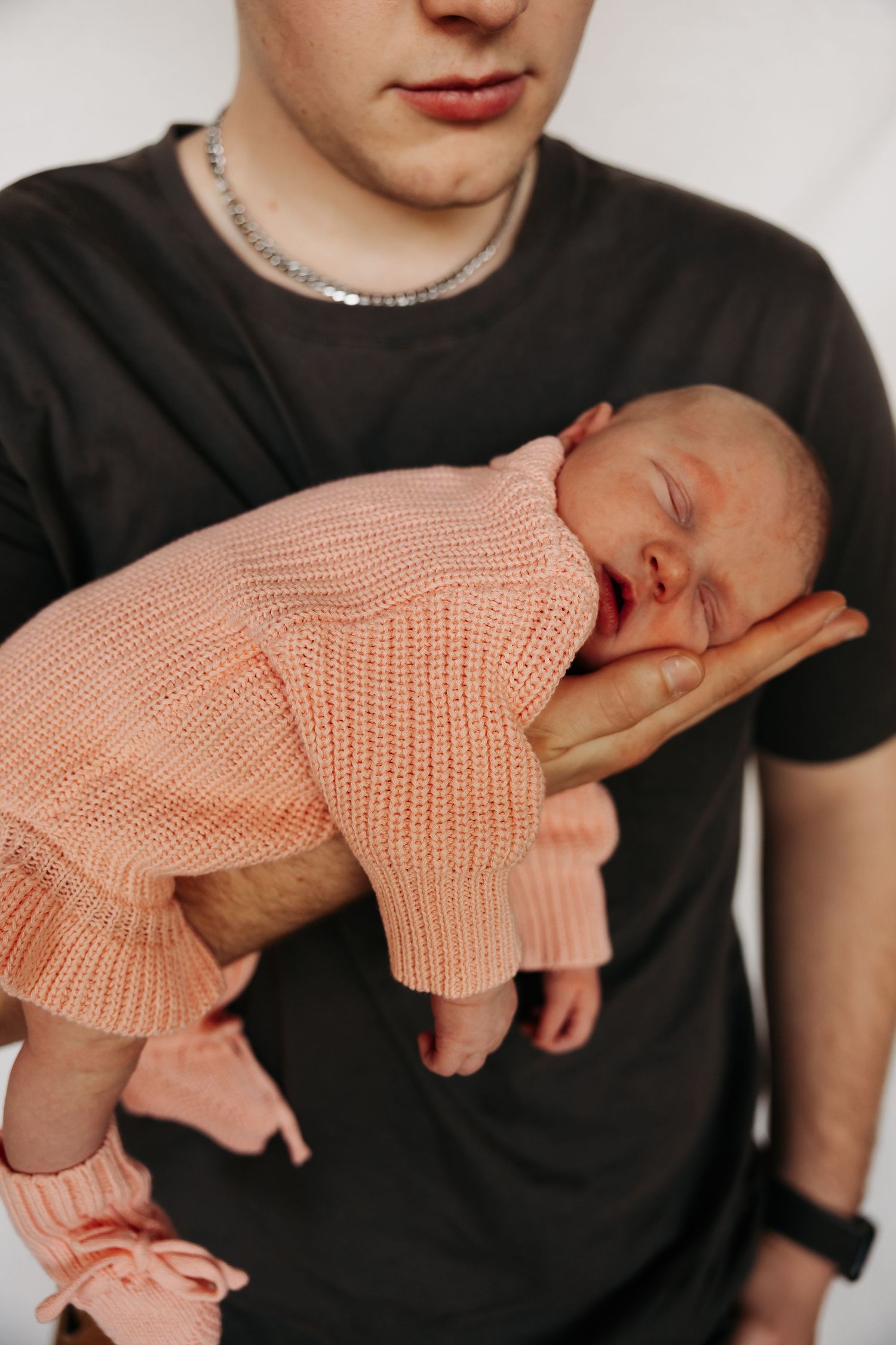 Young father in black t-shirt tenderly holds sleeping newborn baby dressed in pink knit outfit on his hand.