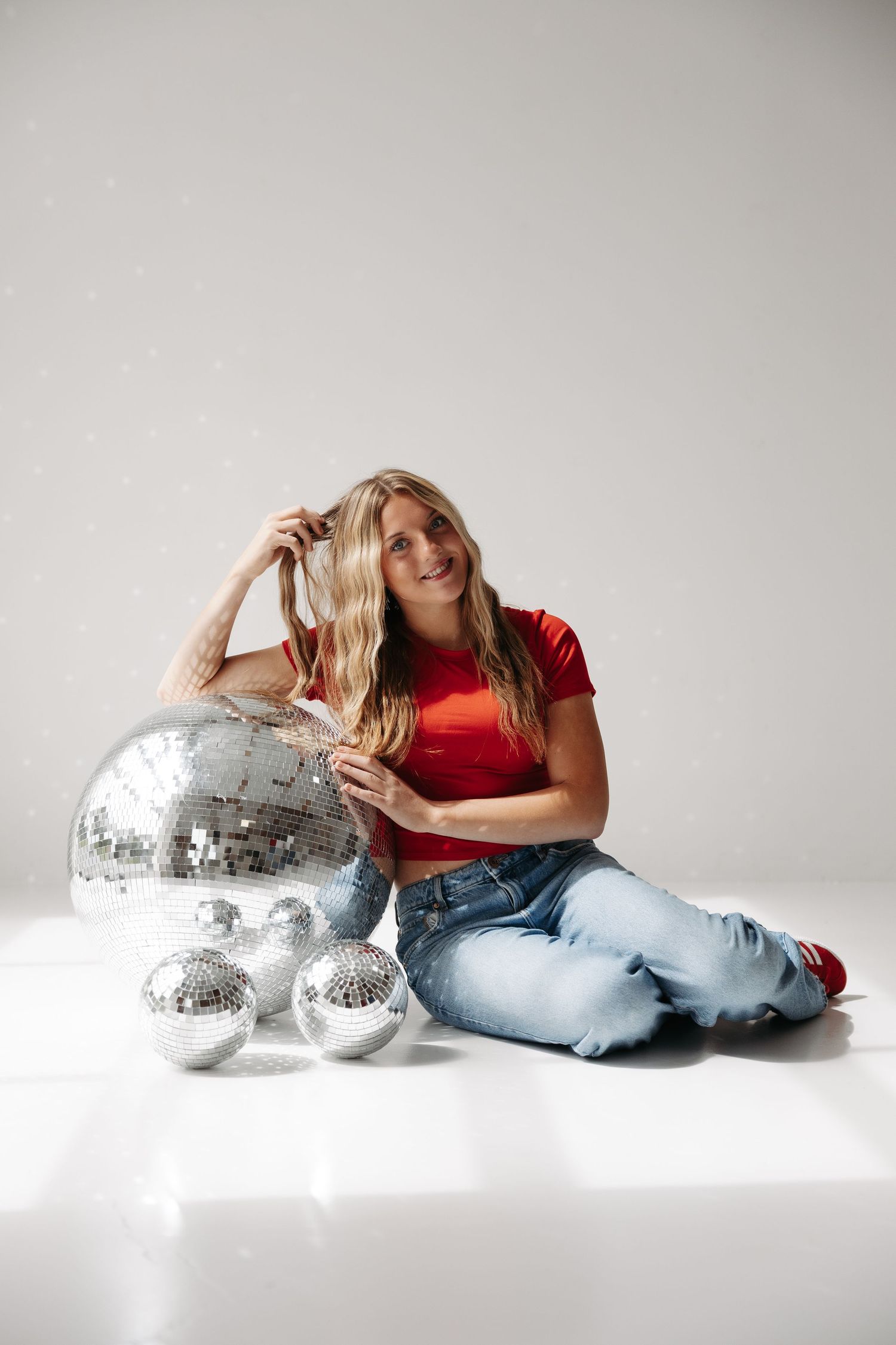 Smiling blonde woman in red top and jeans sitting on white floor with silver disco balls in a studio setting.