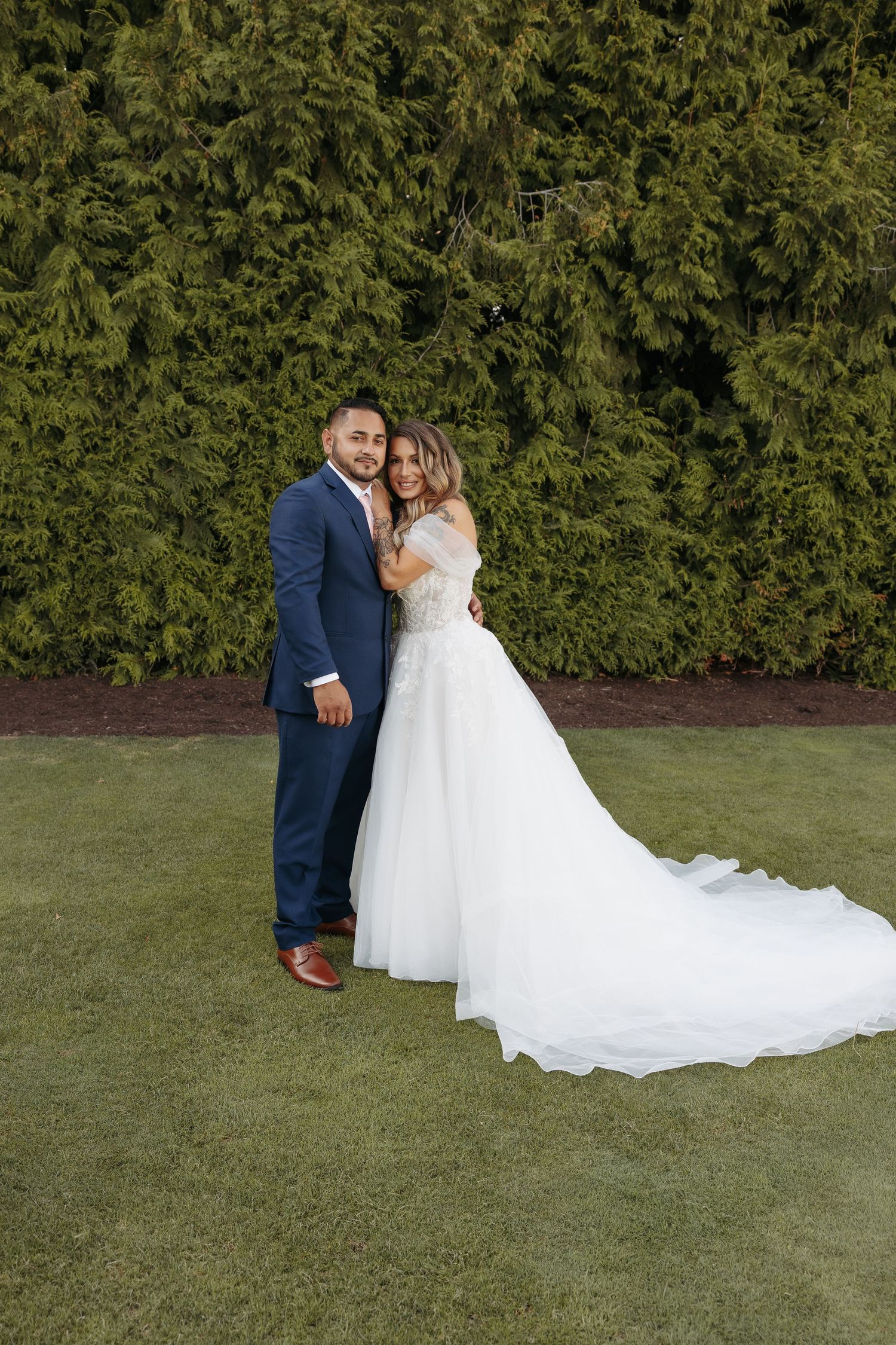A couple in wedding attire poses together on a green lawn with a hedge backdrop.