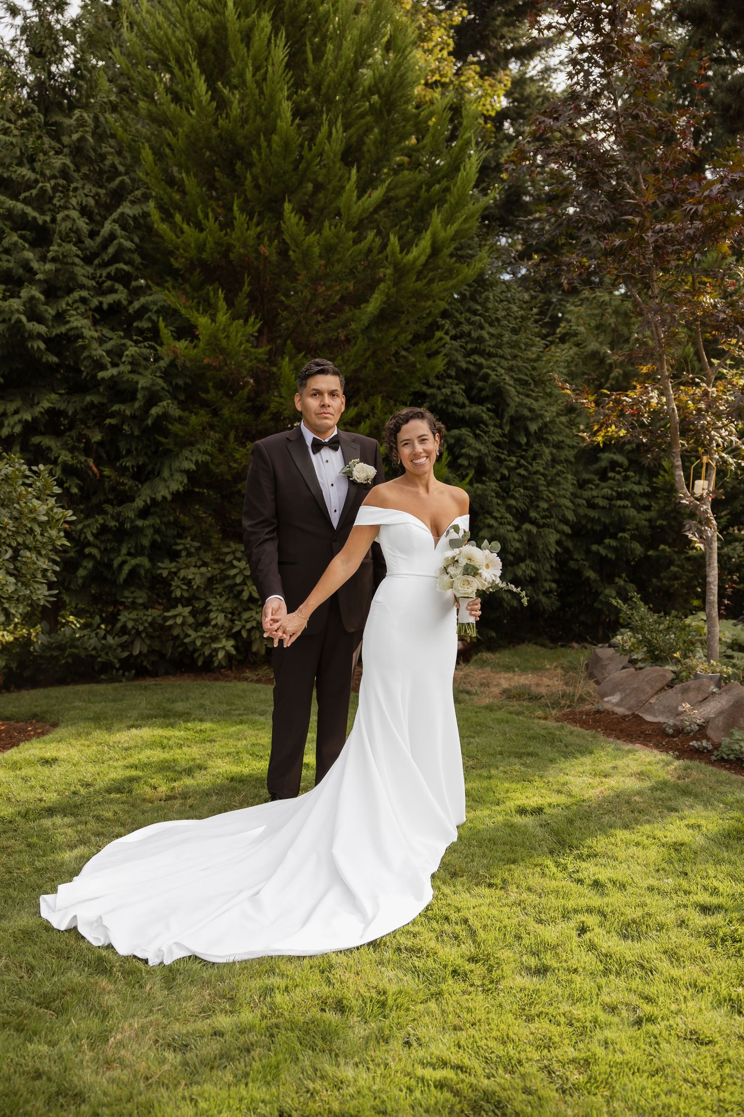 A romantic wedding couple poses on a grassy hill with evergreen trees in the background.
