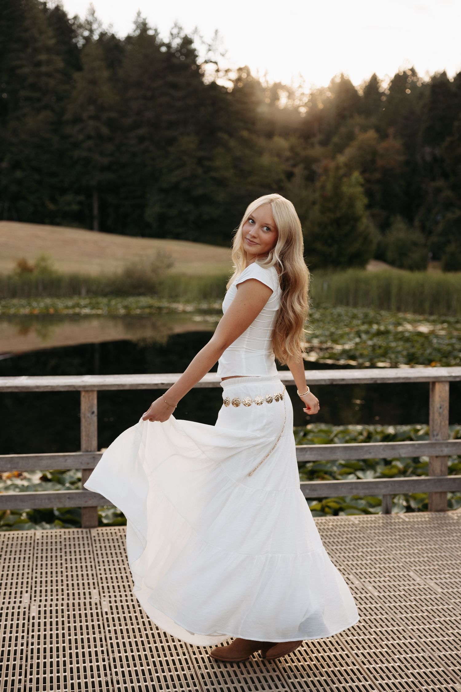 A figure in a flowing white dress twirls gracefully on a wooden dock by a serene lake at sunset.