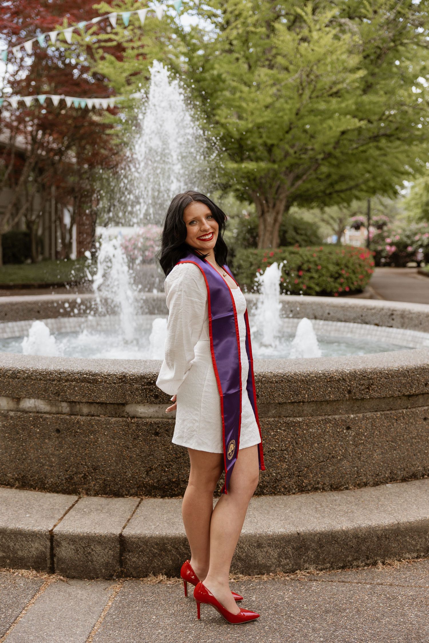 A figure poses in front of a decorative fountain wearing a white dress shirt, blue and red accents, and red high heels.