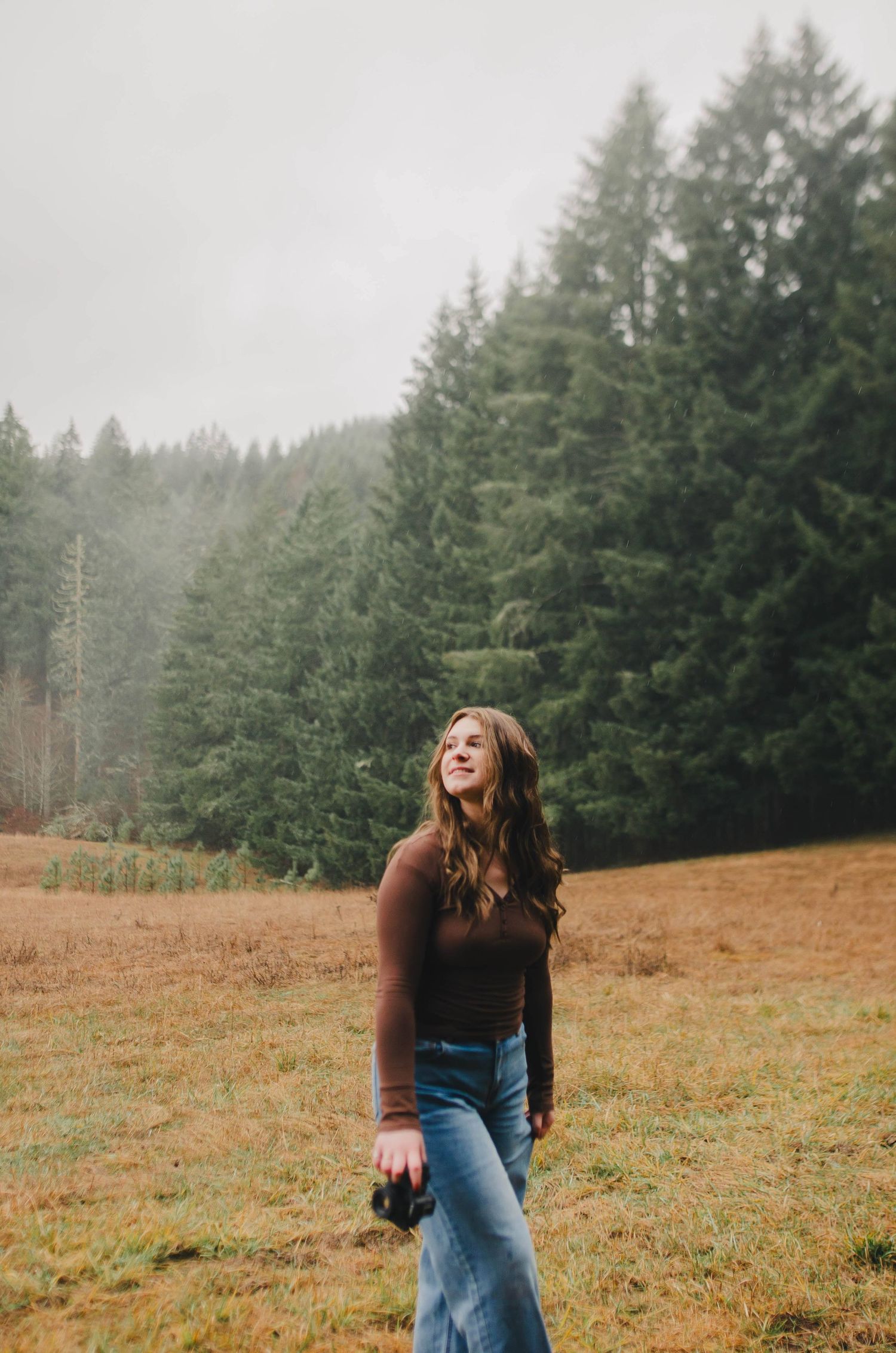 Photographer laughing while taking pictures in autumn field with evergreen forest backdrop.