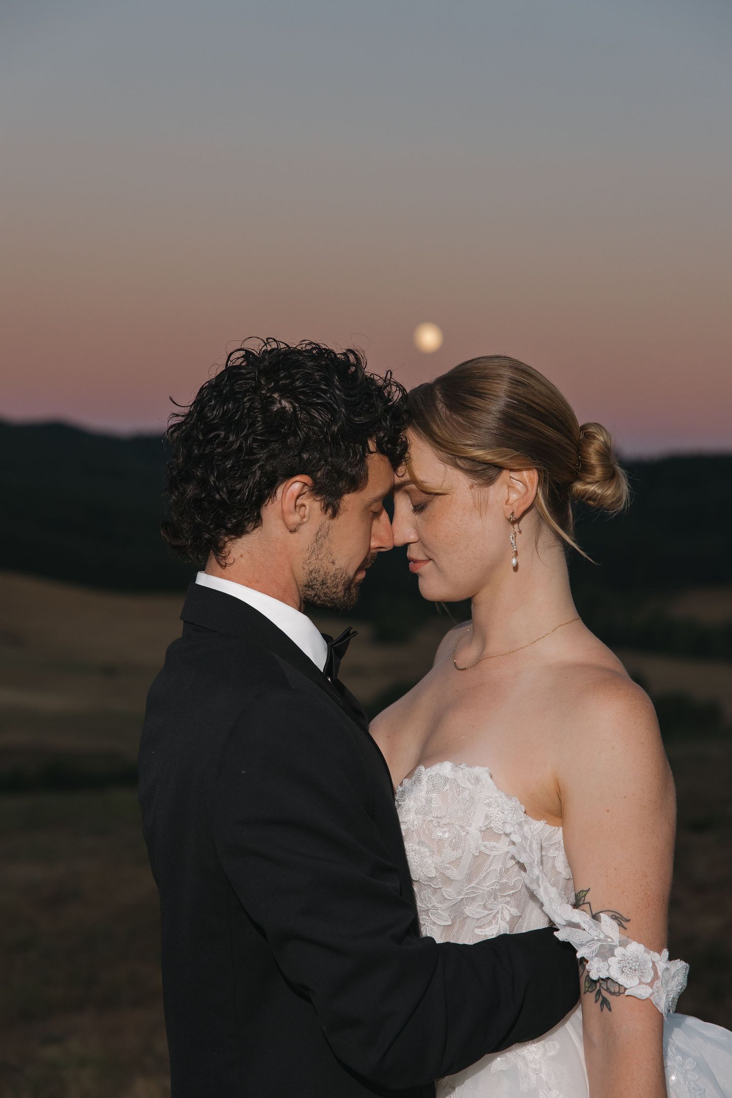 Bride and groom touching foreheads at sunset with full moon rising over rolling hills in romantic wedding portrait.