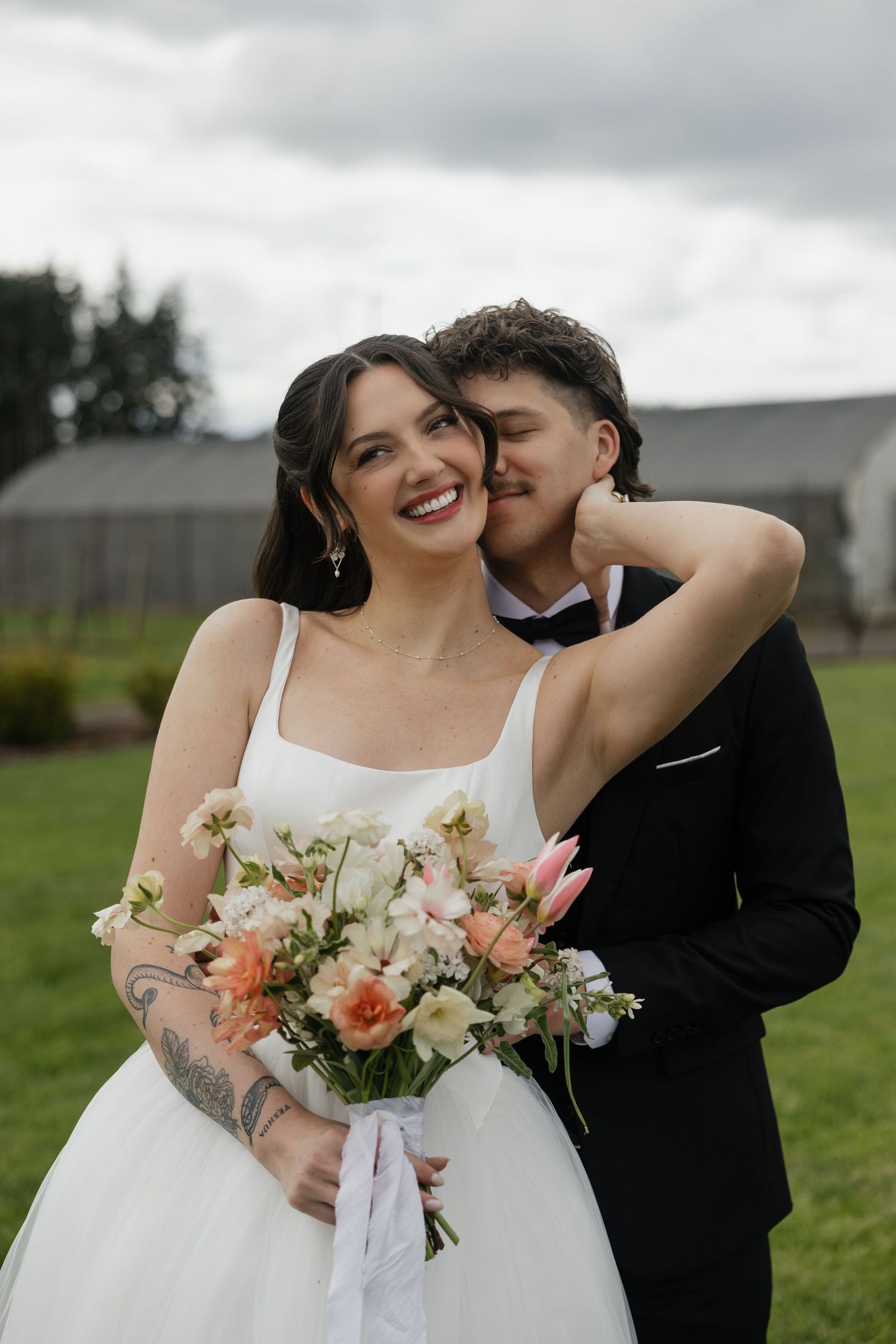 A smiling bride holding a pastel bouquet embraces her groom in a black suit outdoors on their wedding day.