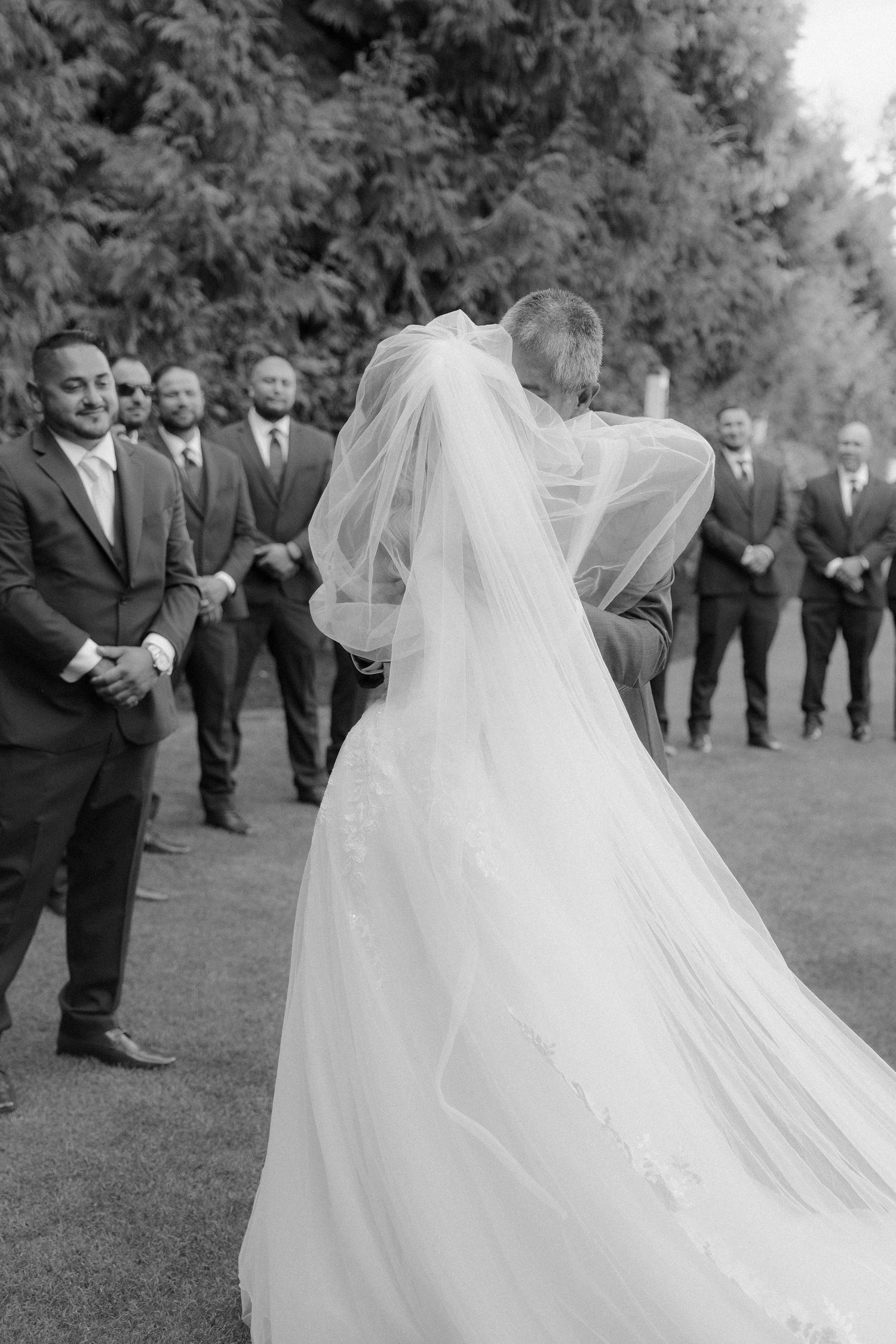 Black and white photo of wedding party on grassy lawn with flowing bridal veil.